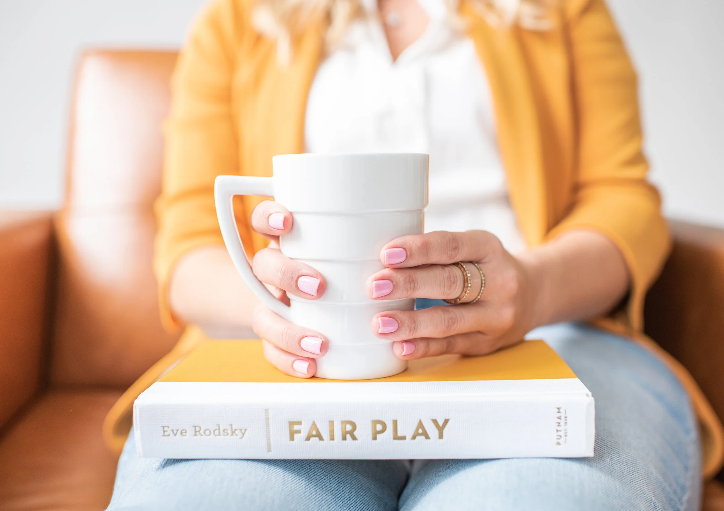 A person in a mustard yellow jacket and white shirt is sitting on a brown couch, holding a white ceramic mug over a yellow and white book titled 'Fair Play' by Eve Rodsky.