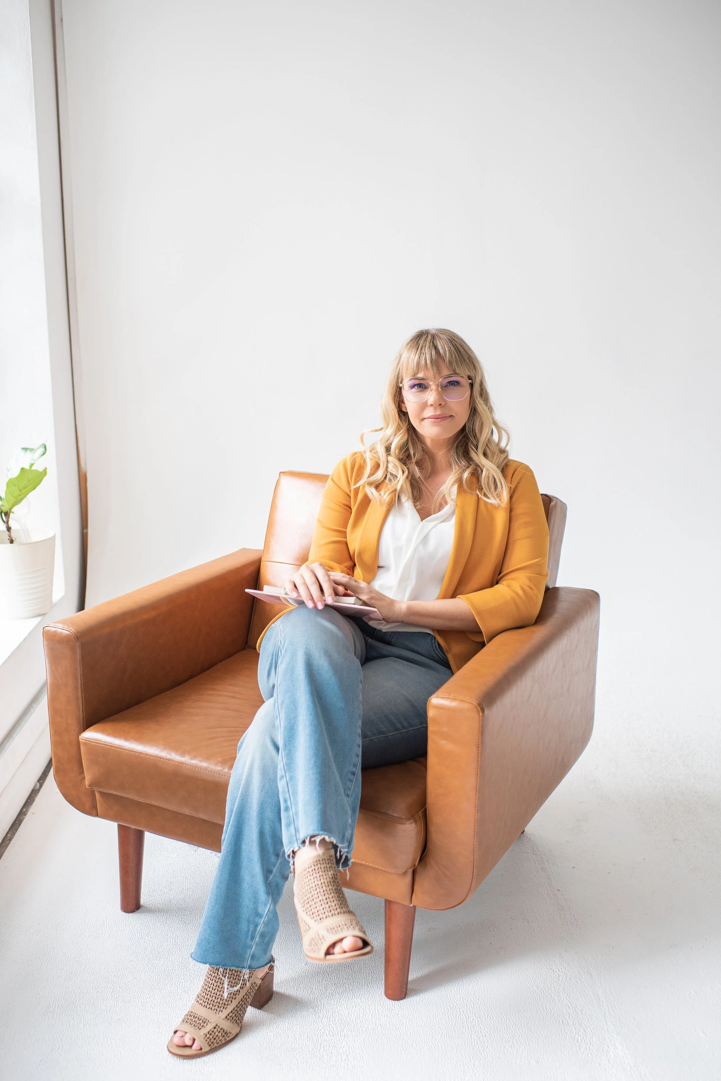 A woman with blond hair and glasses sitting on a brown armchair in front of a white wall, holding a pink notebook and pen, in a bright room with a plant on a windowsill.