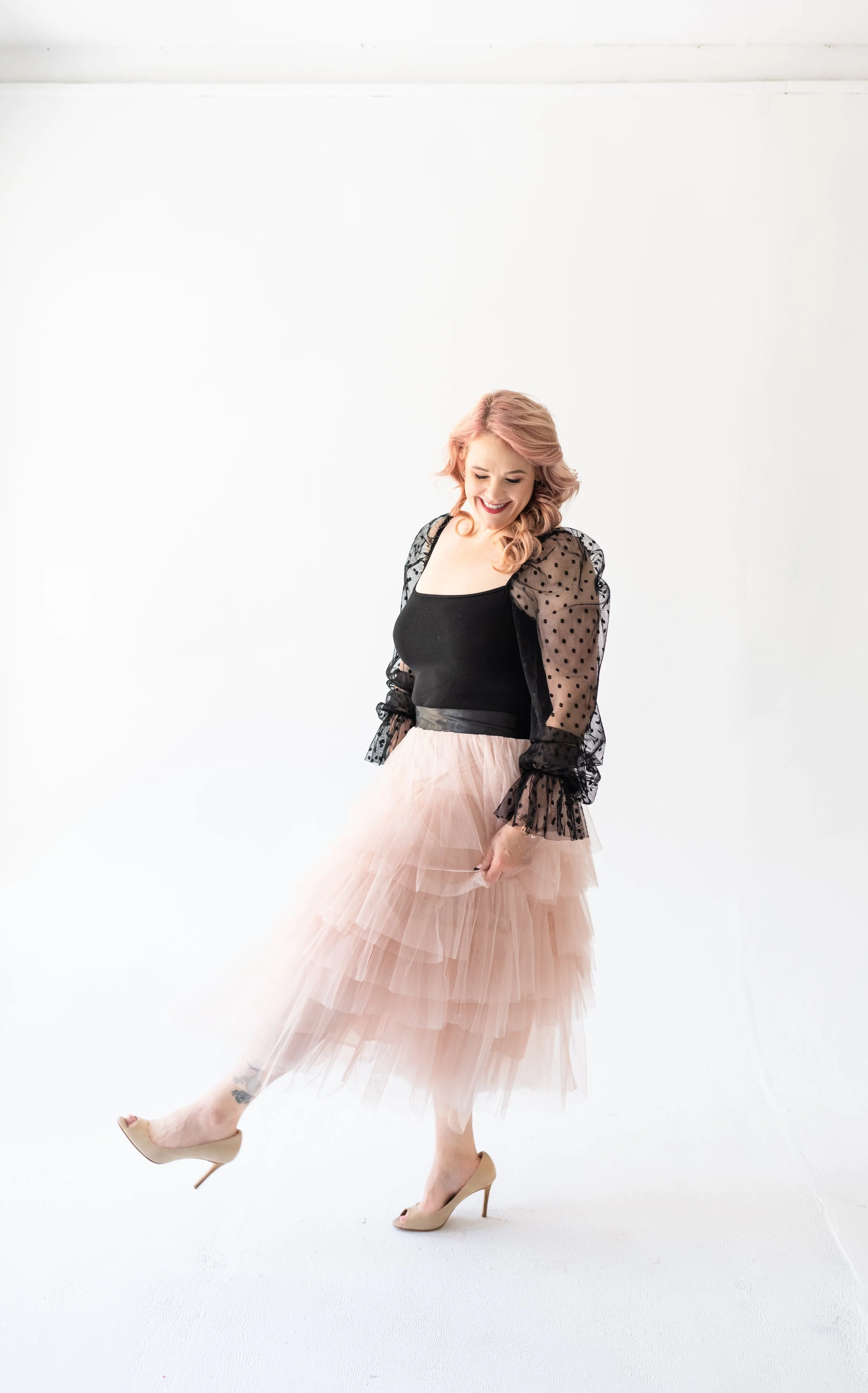 A woman in a black top, pink tulle skirt, black polka-dot sheer jacket, and nude high heels standing against a plain white background, smiling and looking downward.