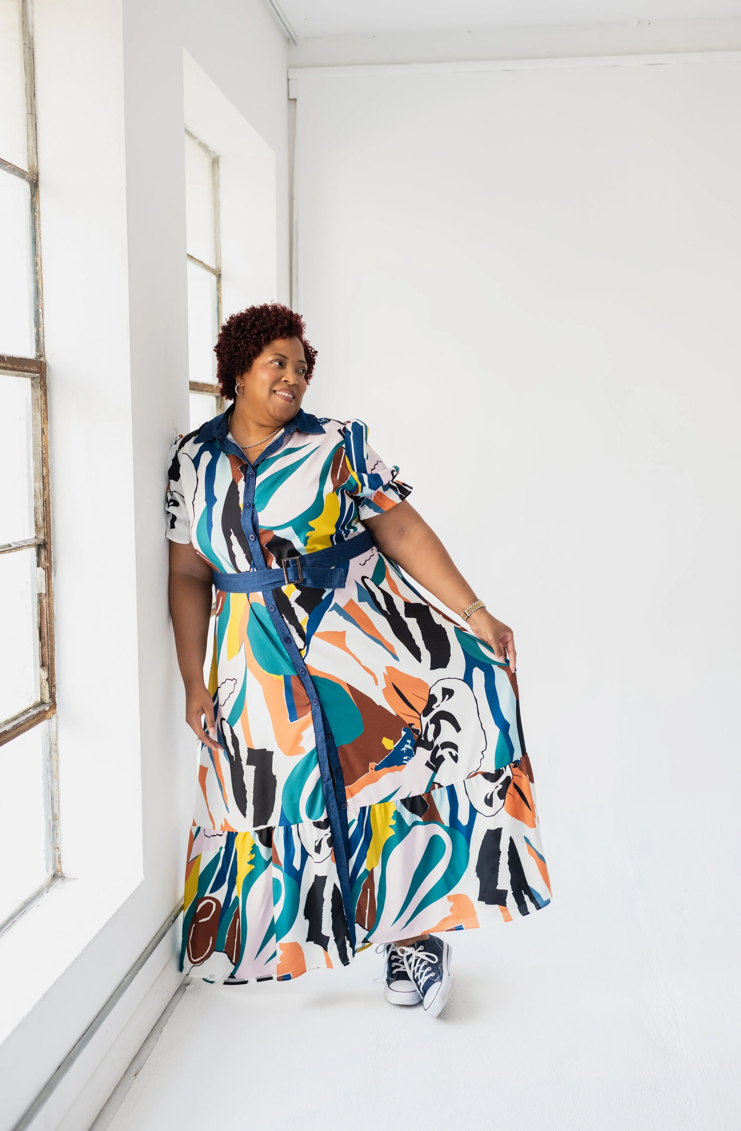Woman in a colorful abstract patterned dress leaning against a white wall by a window.