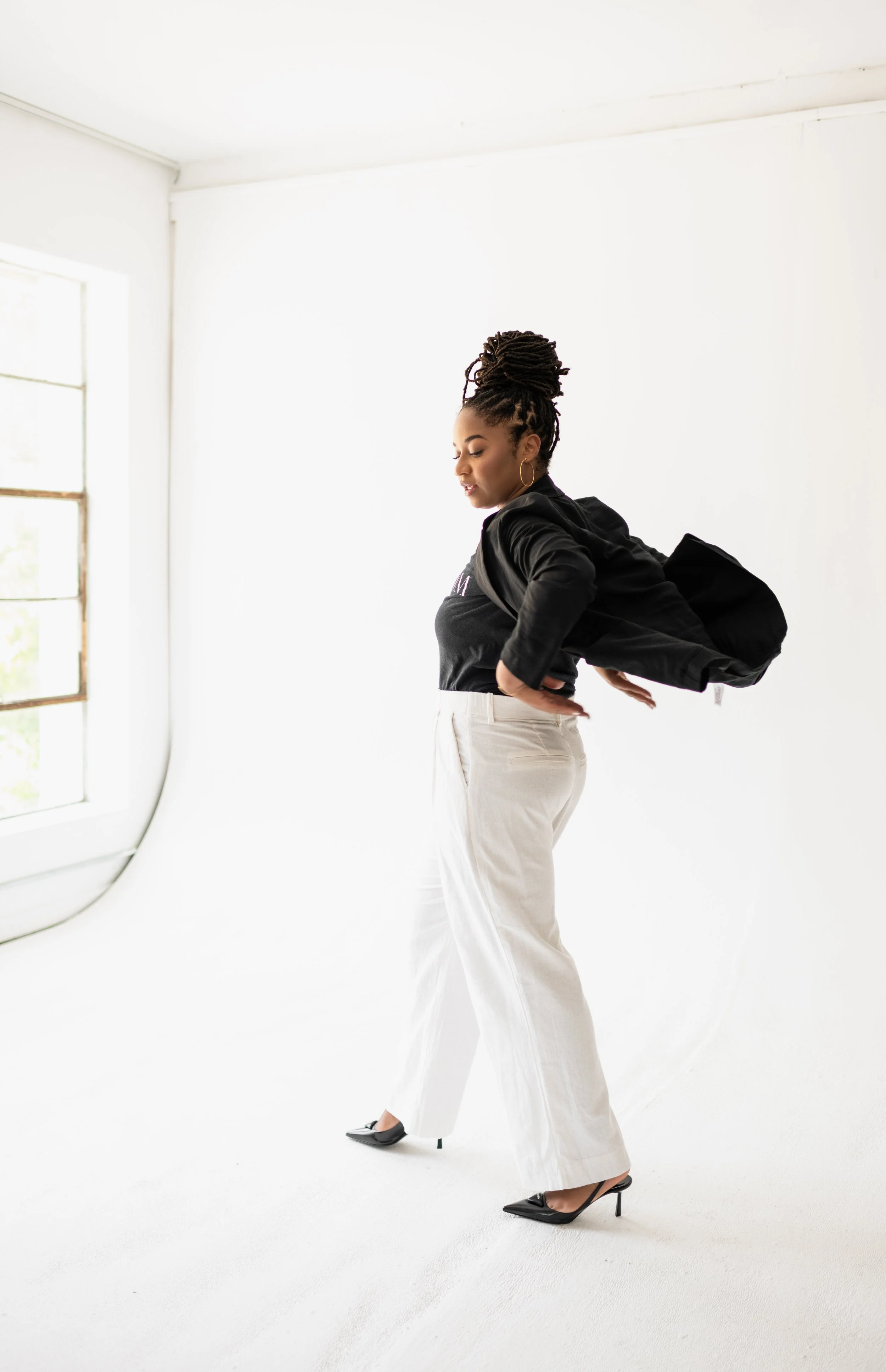 A woman standing in a bright room with white walls, adjusting her black jacket while wearing white pants and black high heels.