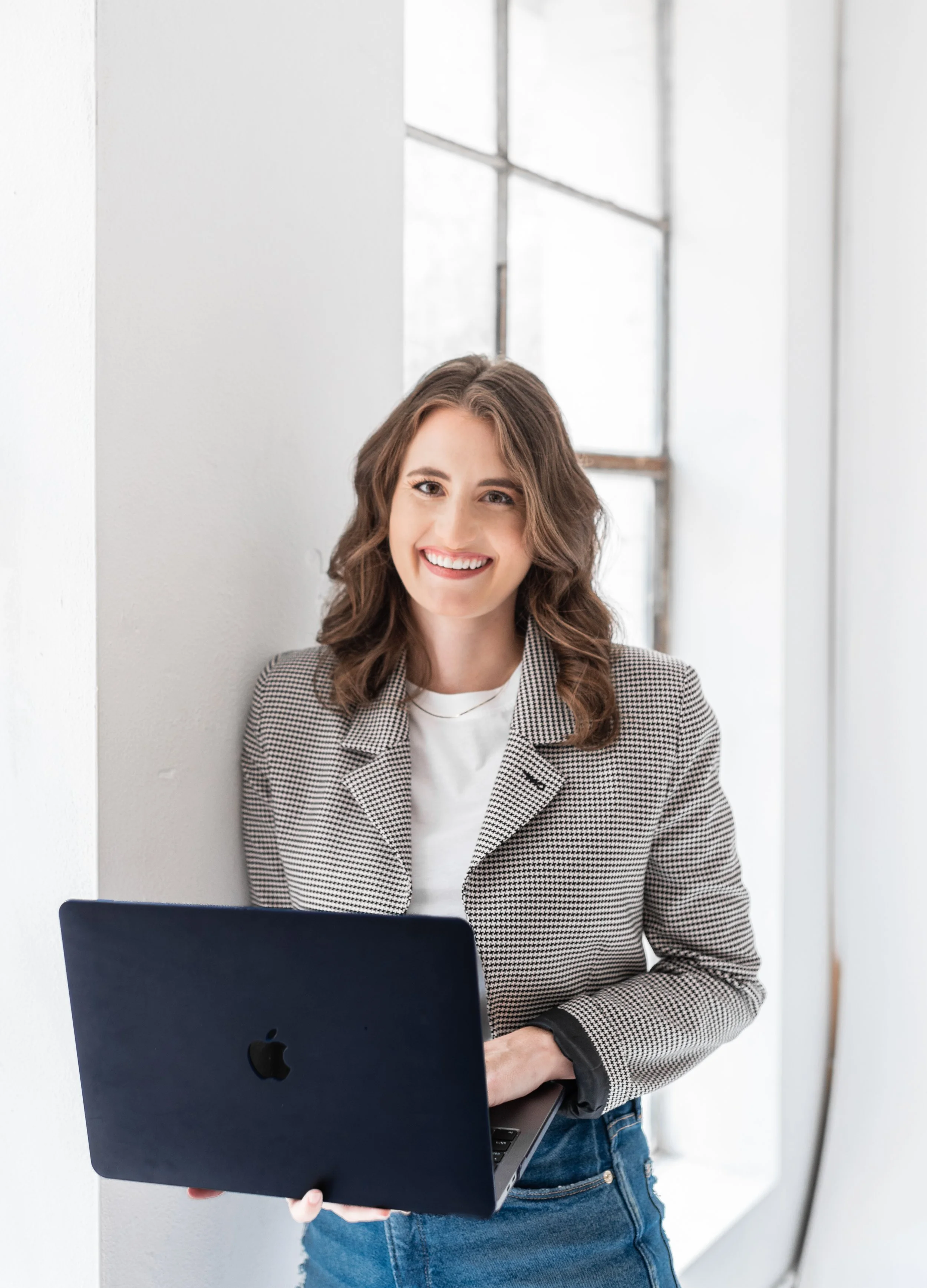 A woman with wavy brown hair, wearing a checkered blazer and white shirt, smiling and holding a black Apple MacBook laptop in a bright room near a large window.