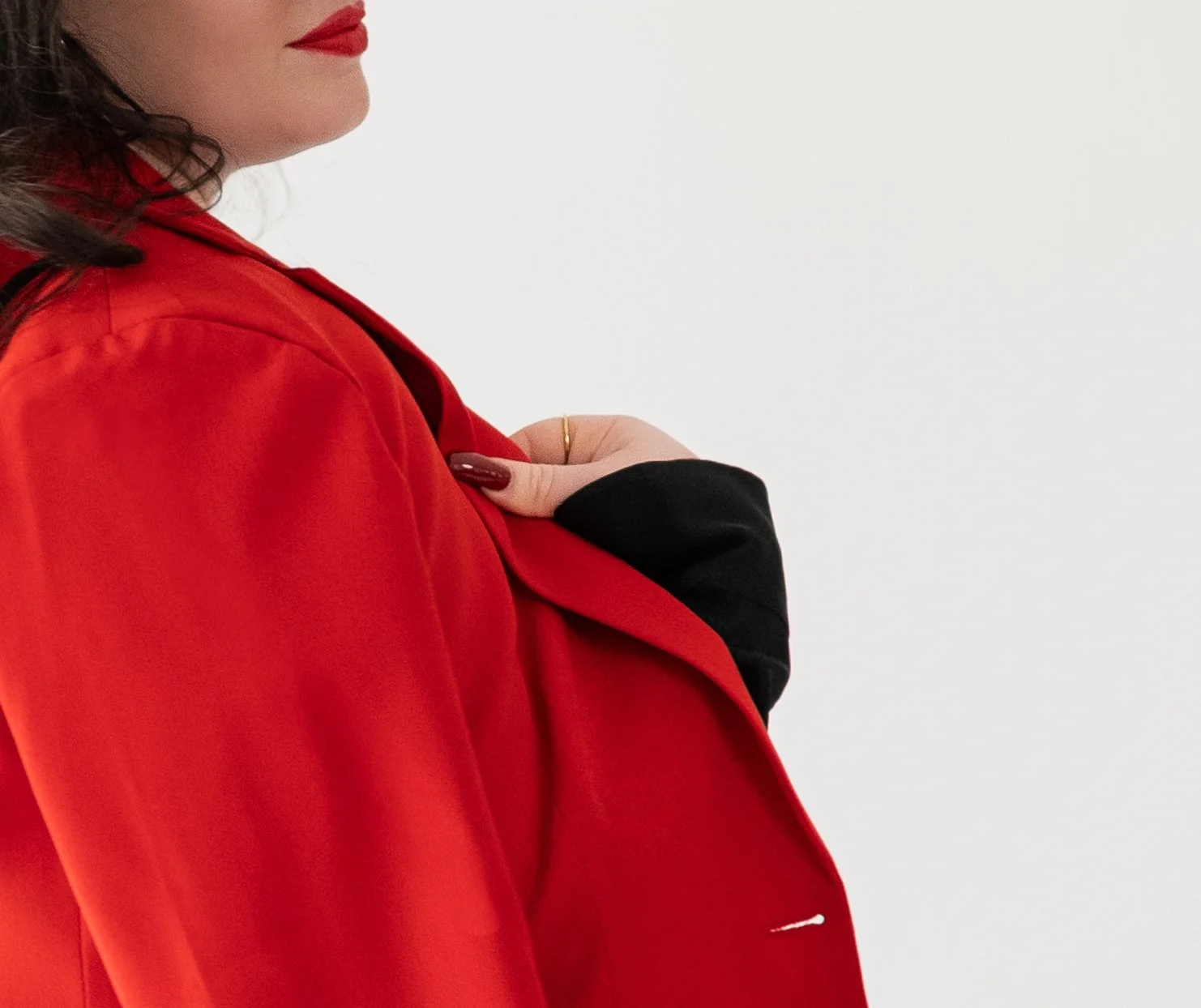 A woman in a red blazer with dark lipstick and a gold ring on her hand holding her blazer closed, against a plain white background.