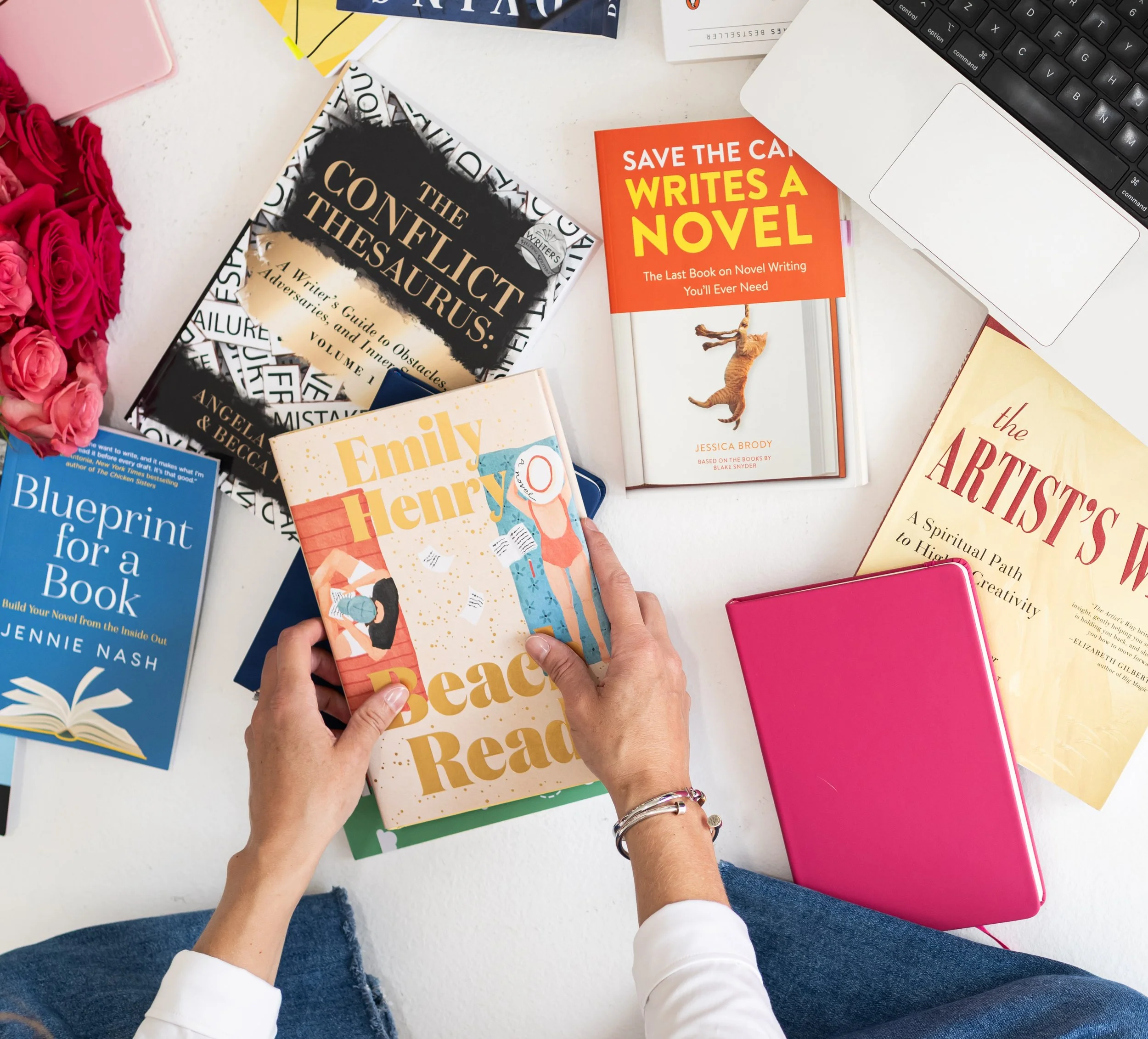 A person holding a book titled "Beach Read" by Emily Henry. Several other books, a pink notebook, a laptop, and a bouquet of pink roses are on the white table.