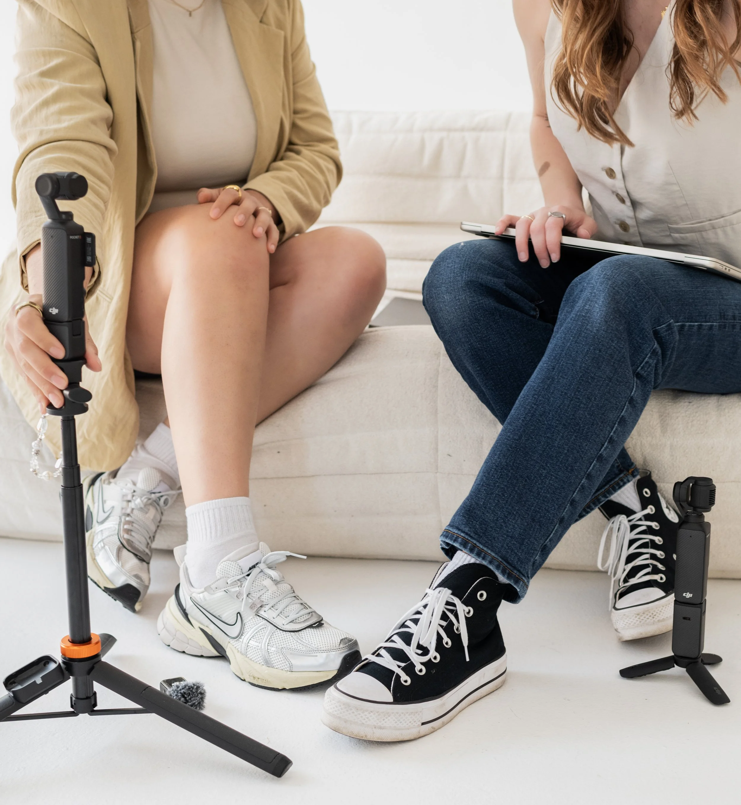 Two women sitting on a cream-colored sofa having a conversation, with recording equipment and a clipboard nearby.