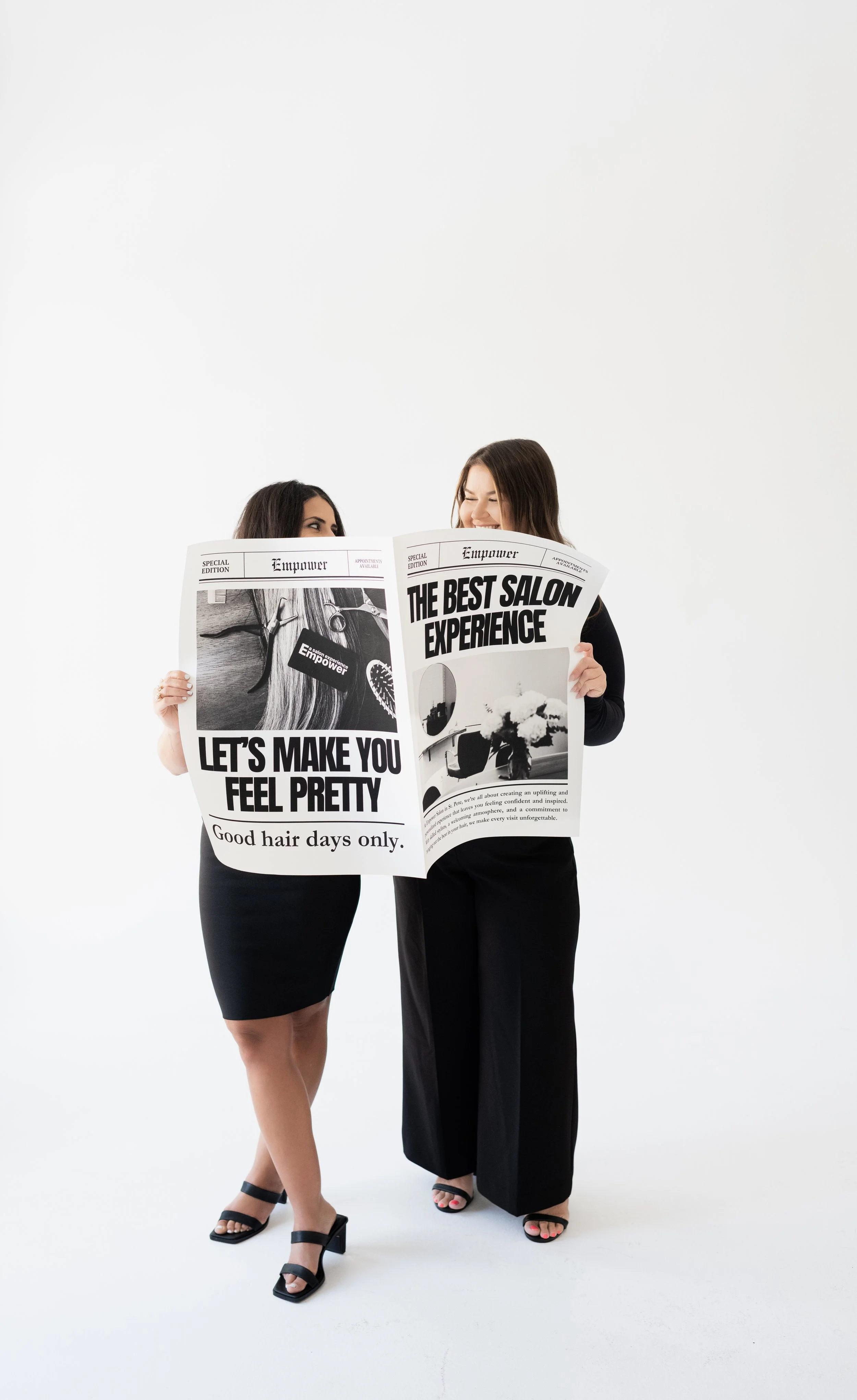 Two women stand on a white background, holding a large newspaper and smiling. One woman wears a black dress, and the other wears black wide-leg pants and a black top. They appear happy and engaged with each other.
