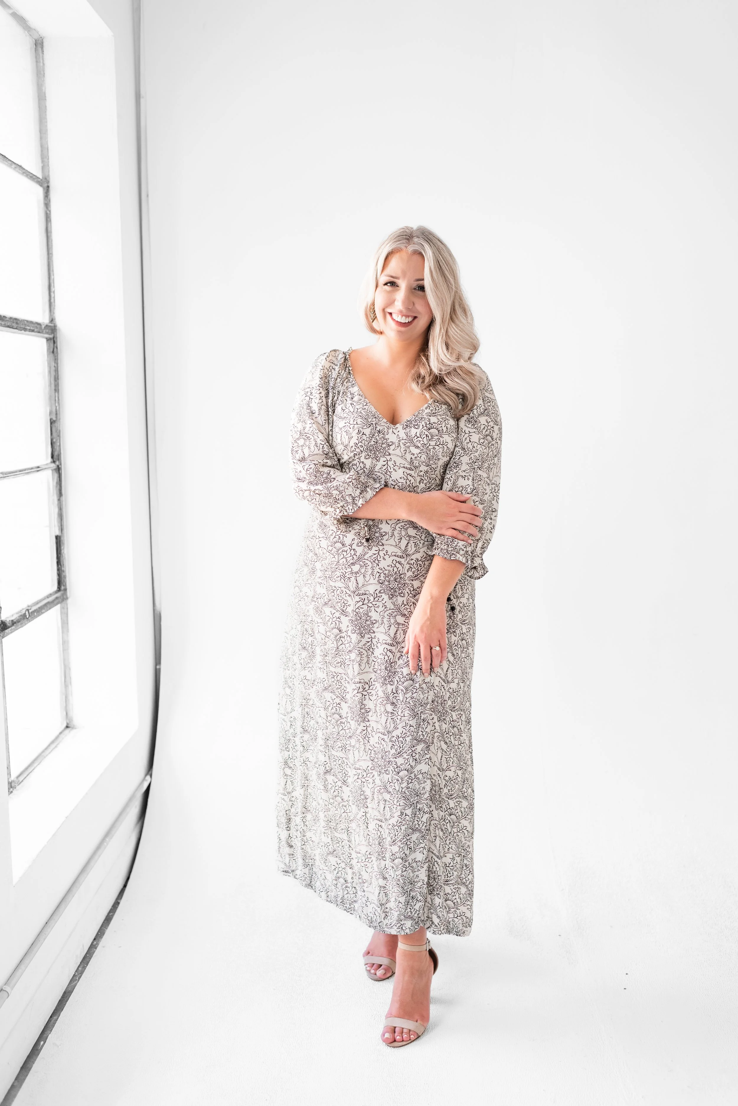 A smiling woman with blonde hair in a long, floral, beige dress stands in a bright white studio with a large window on the left.