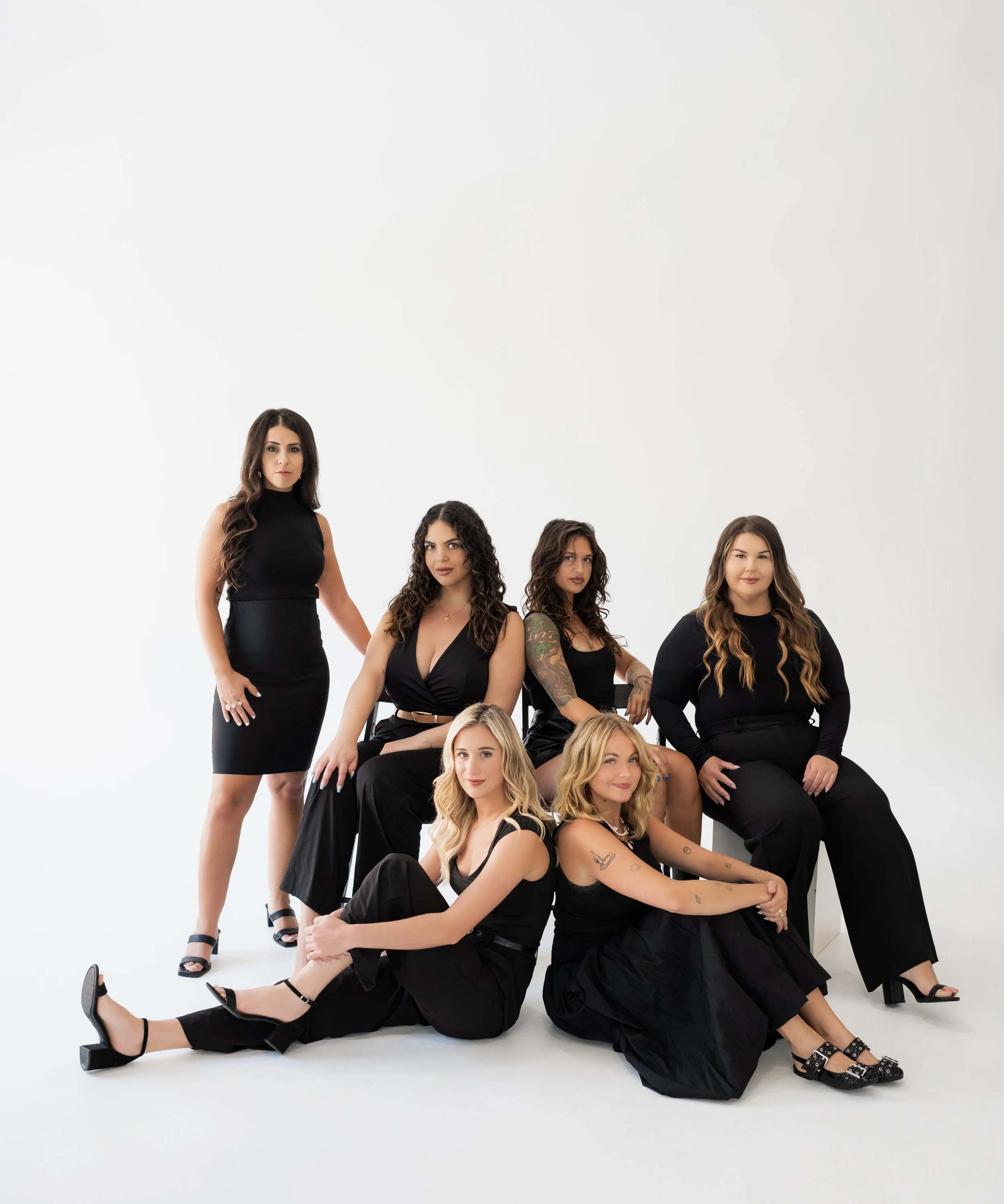 A group of six women dressed in black posing in a studio with a white background.