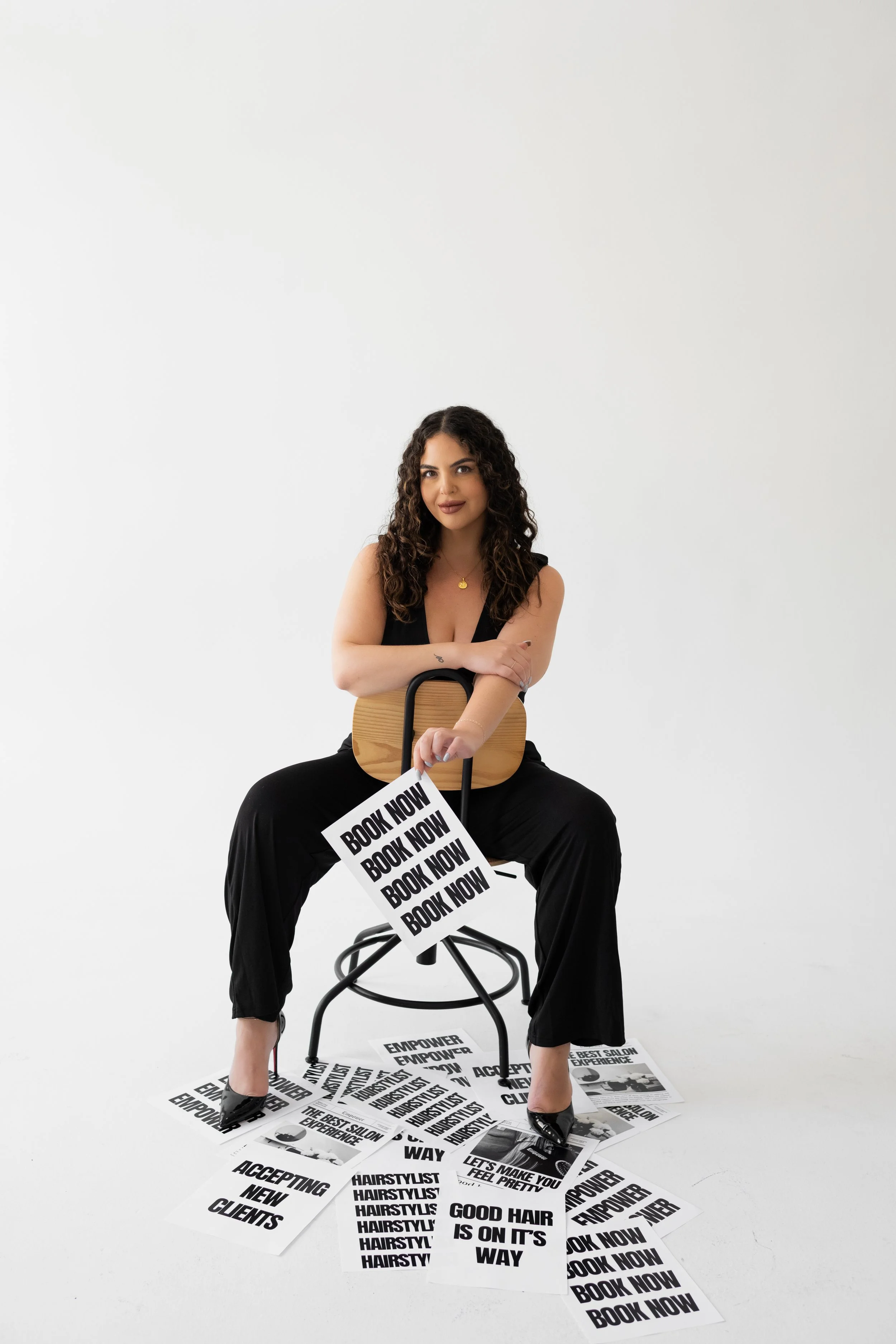 A woman with dark curly hair sitting on a chair in a white studio, surrounded by printed signs with promotional messages, holding a sign that says 'BOOK NOW.'