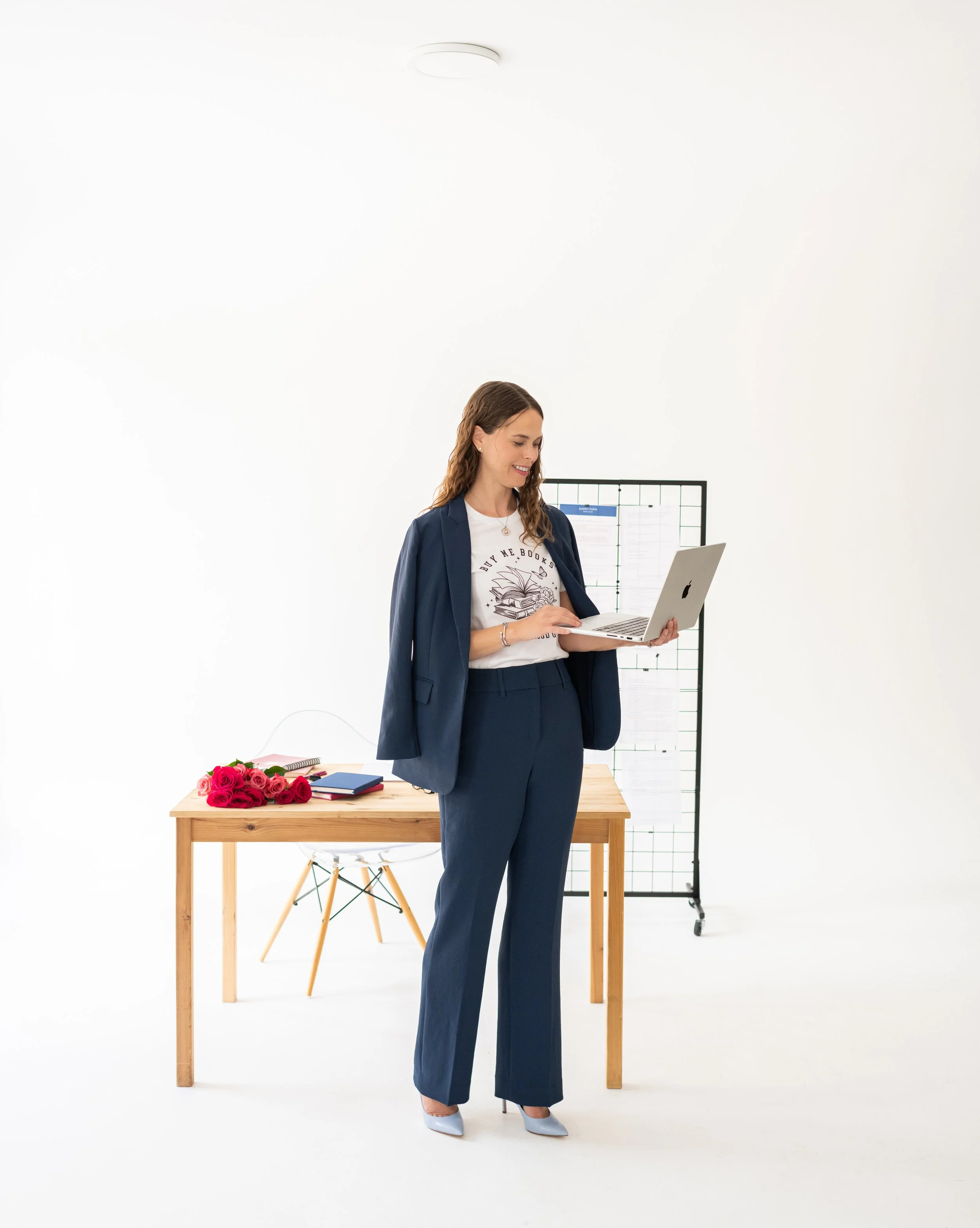 A woman in a navy suit standing at a desk, working on a laptop, with a bouquet of red roses and notebooks on the desk, and a whiteboard in the background, in a minimalist office setting.