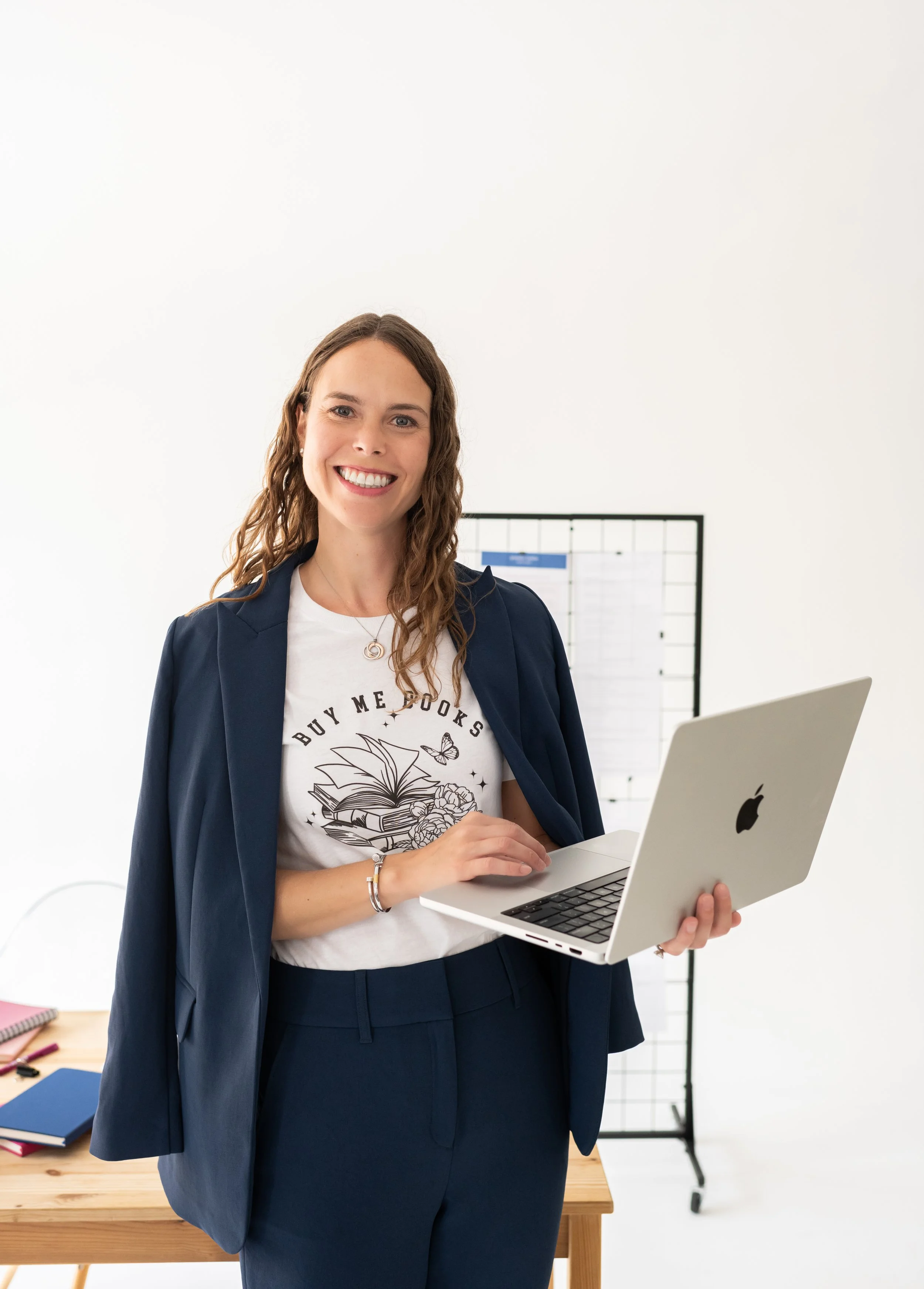 A smiling woman in business attire standing in an office, holding a silver laptop with an Apple logo, with a white wall and a black grid in the background.