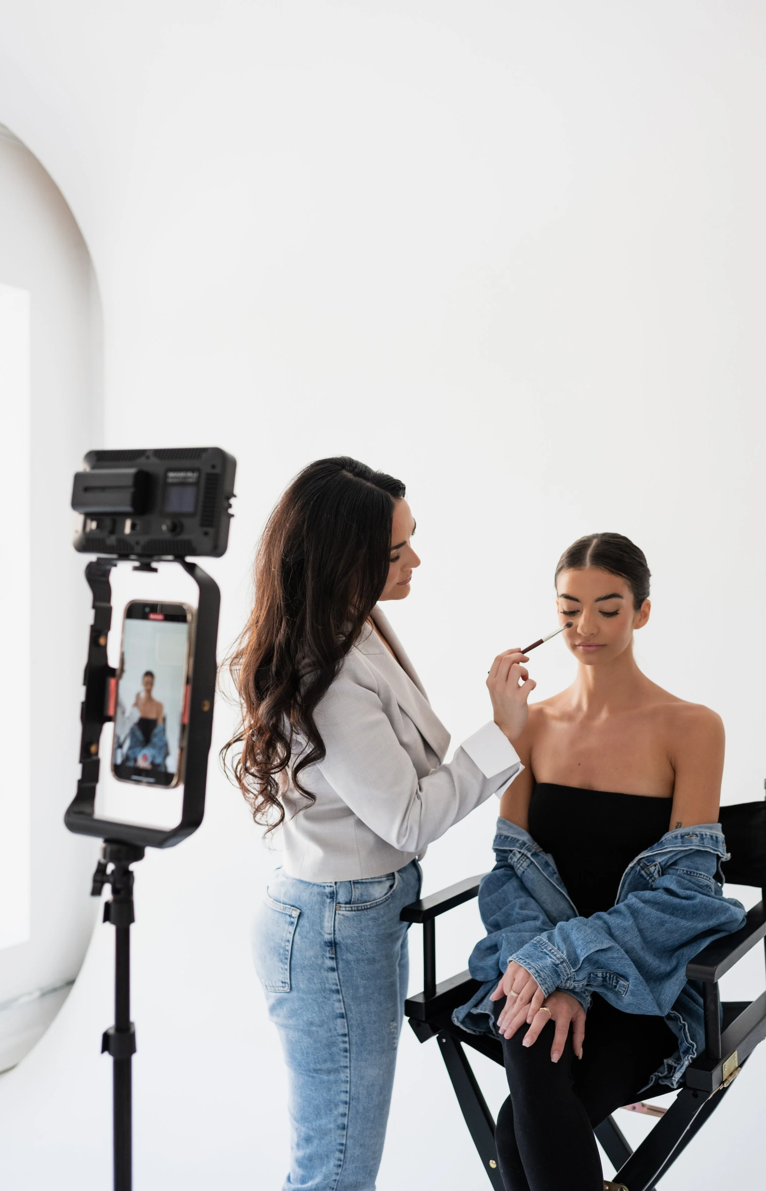 Makeup artist applying makeup to a seated woman in a photography studio.