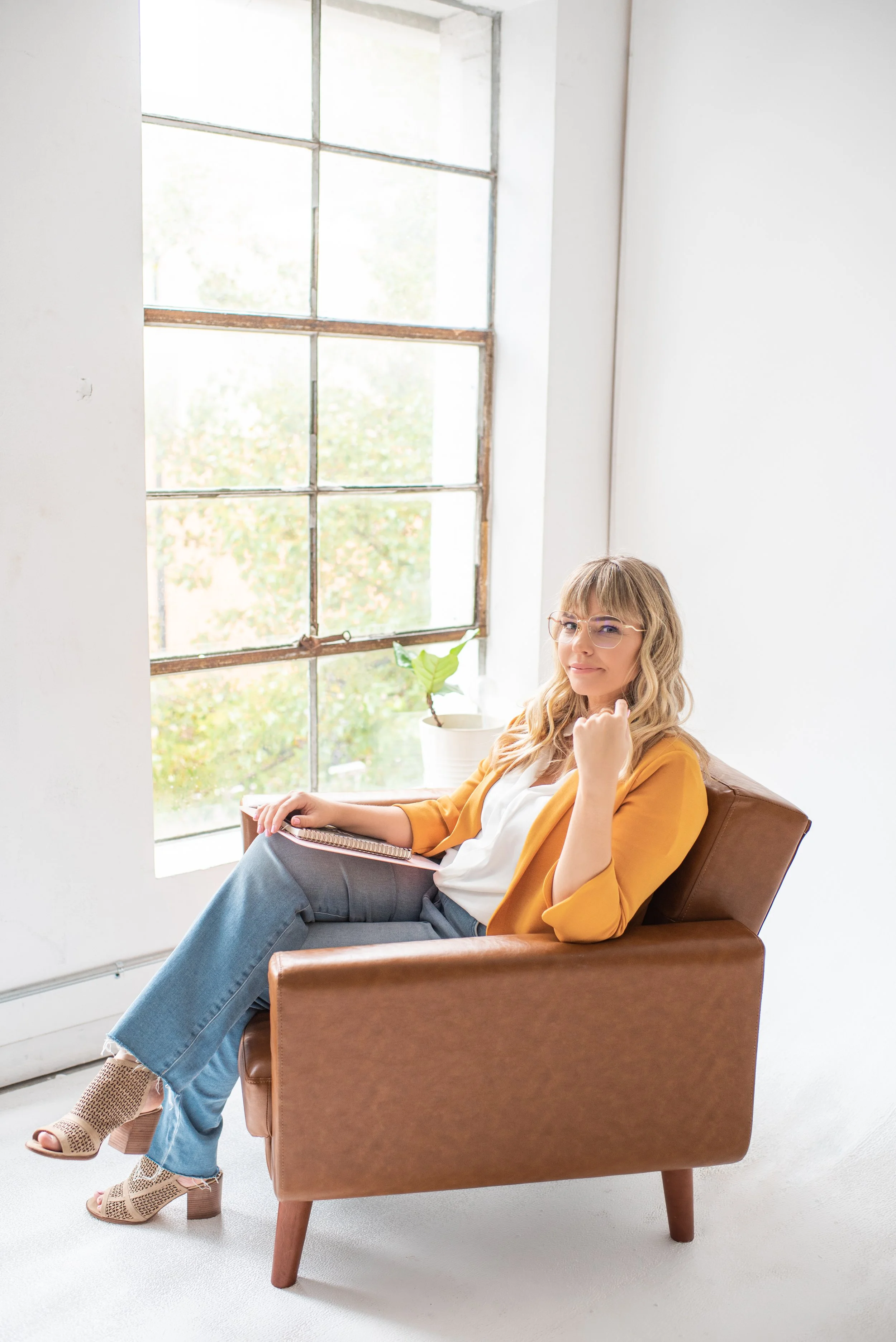 Woman with glasses sitting in a brown armchair by a large window with a potted plant nearby.
