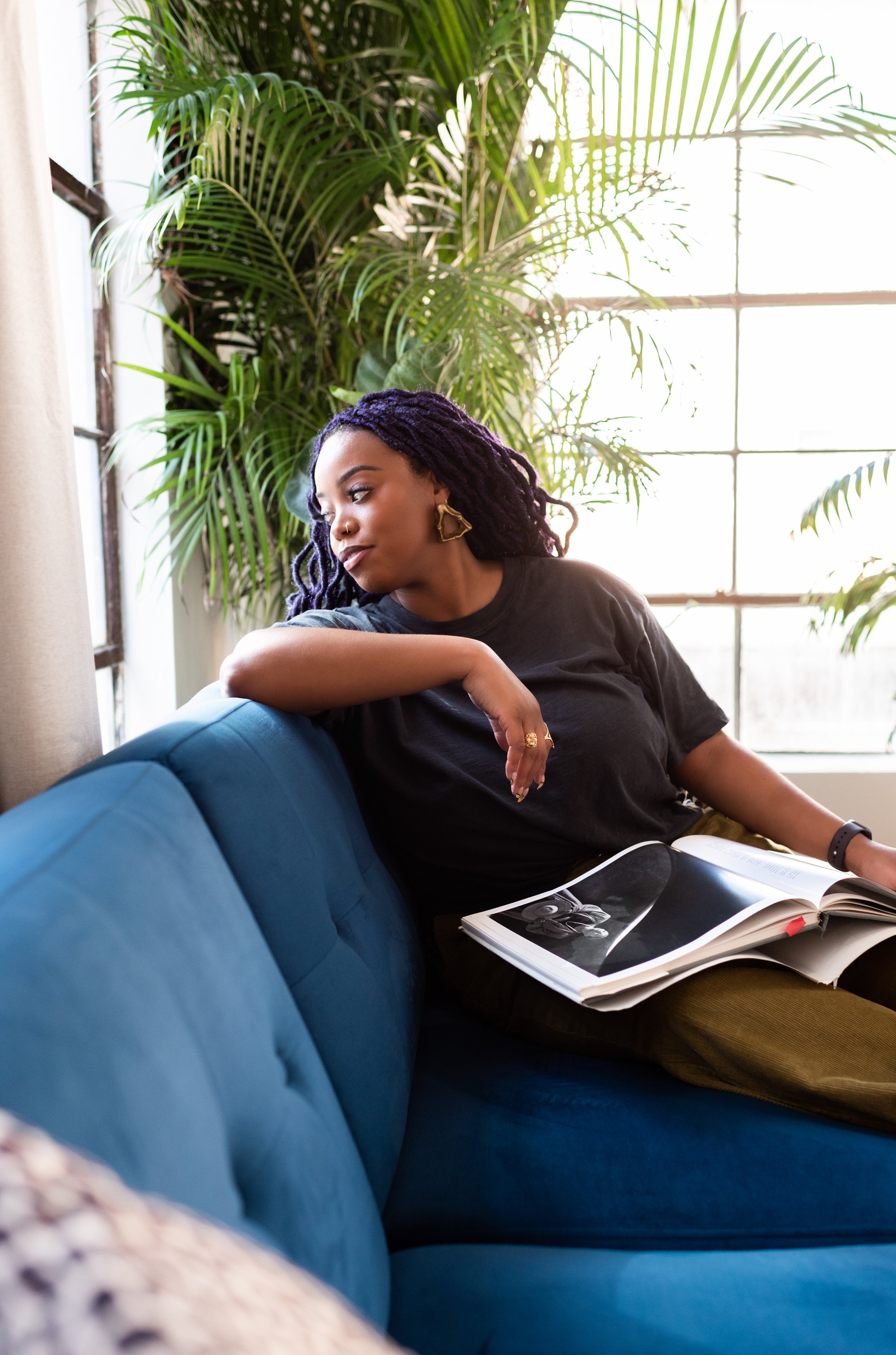 A woman with purple dreadlocks wearing a black t-shirt and gold jewelry sitting on a blue couch, reading a magazine, with large green plants and a window in the background.