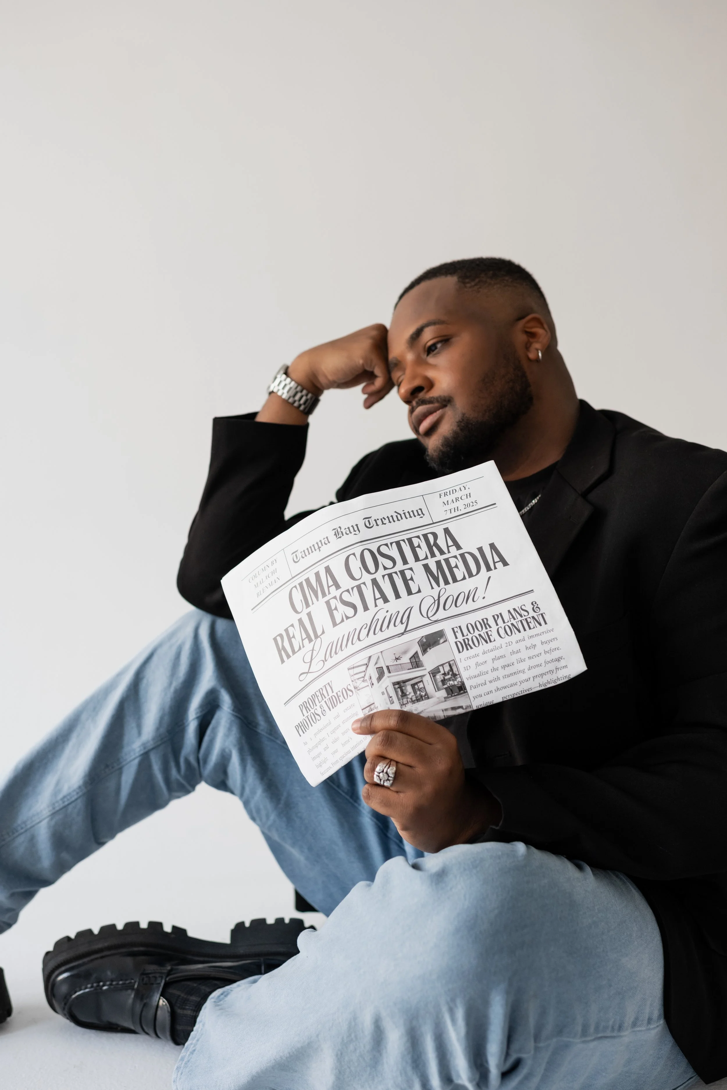A man sitting on the floor and reading a newspaper titled 'Cima Coster Real Estate Media' with a headline 'Launching Soon!'. He is wearing a black blazer, light blue jeans, black boots, and a silver watch. He has a thoughtful expression, a beard, and