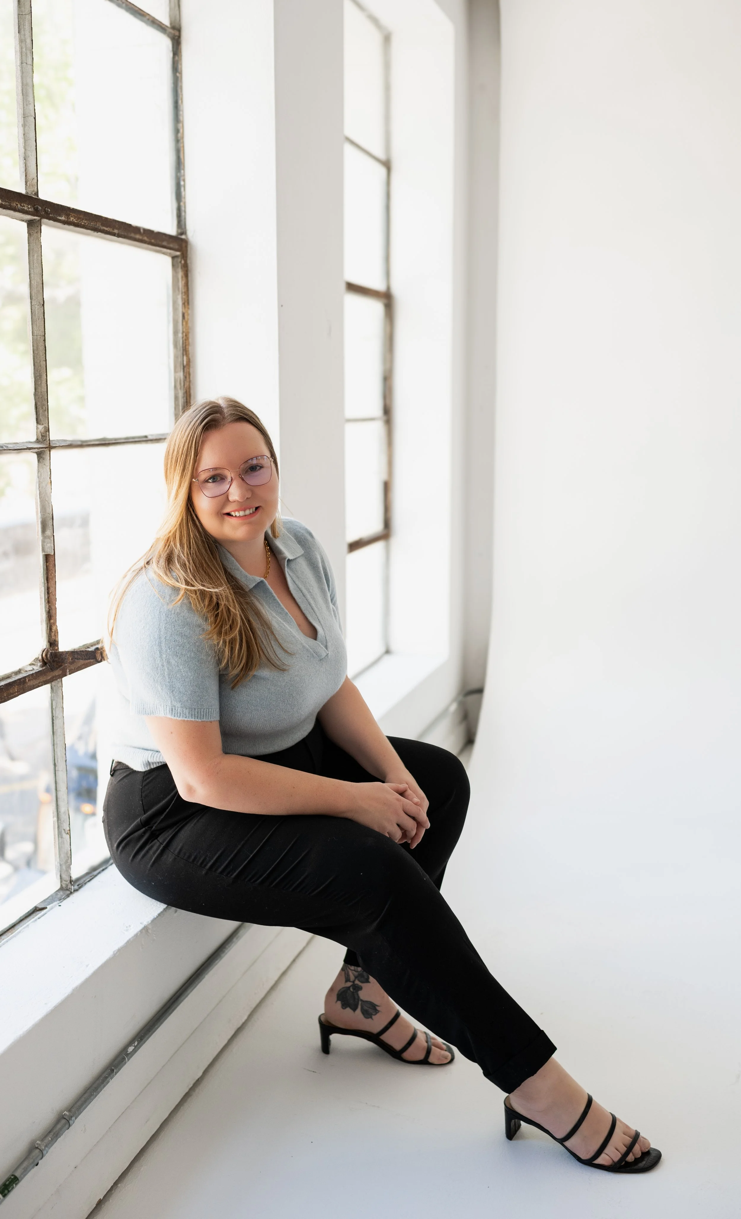 A woman with long hair, glasses, and a tattoo on her ankle, sitting on a window ledge in a bright, minimalistic room, smiling at the camera.
