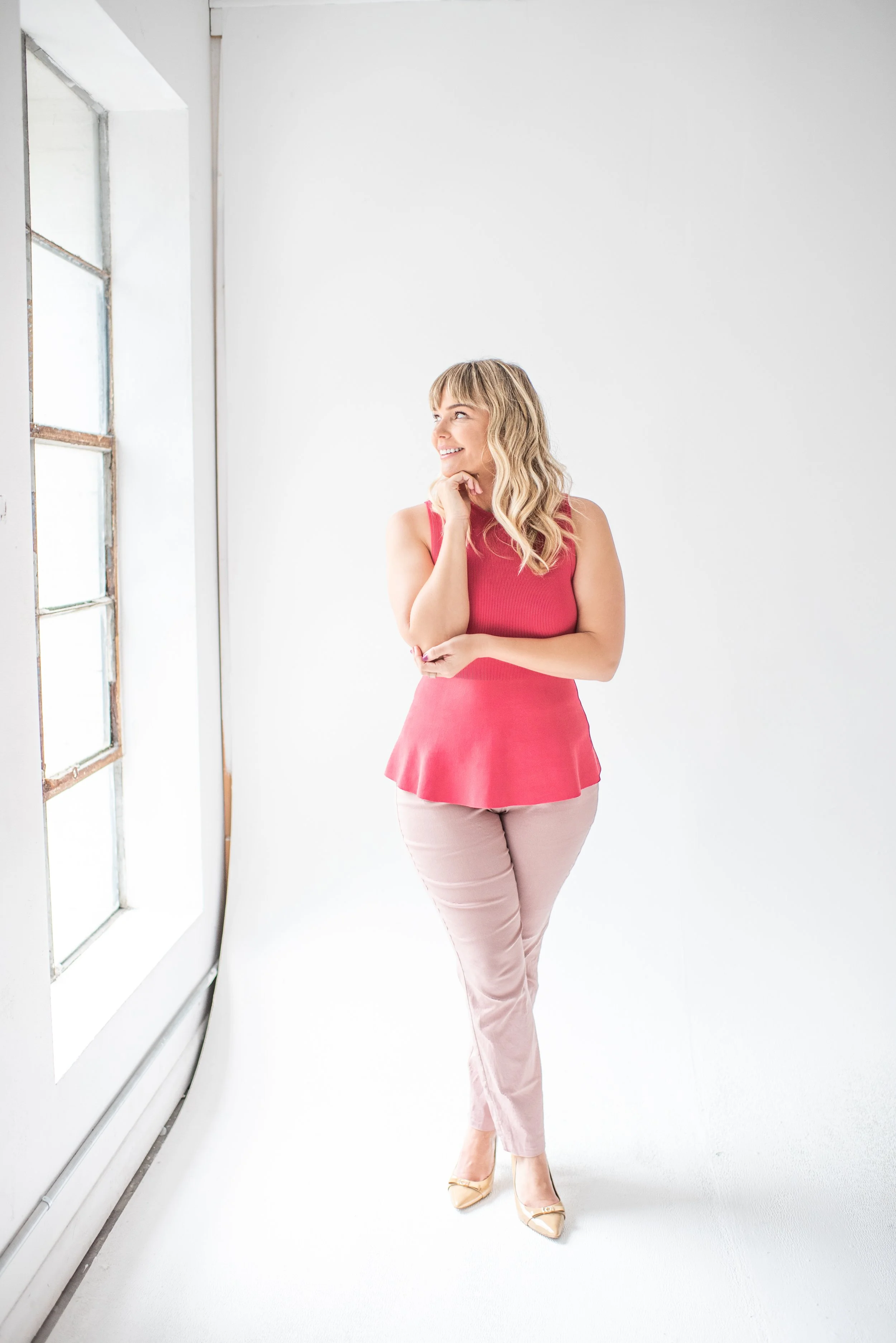 A woman in a pink sleeveless top and light pink pants standing in a bright room with large windows, smiling and looking to her left.