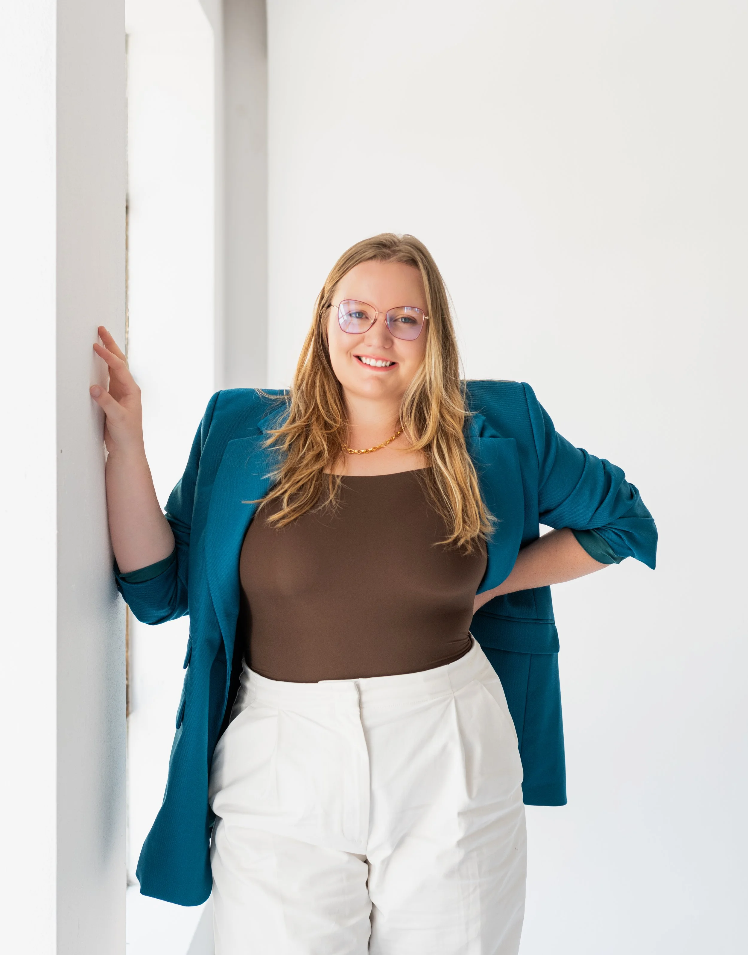 A confident woman in glasses and a blue blazer standing against a white background, smiling at the camera.