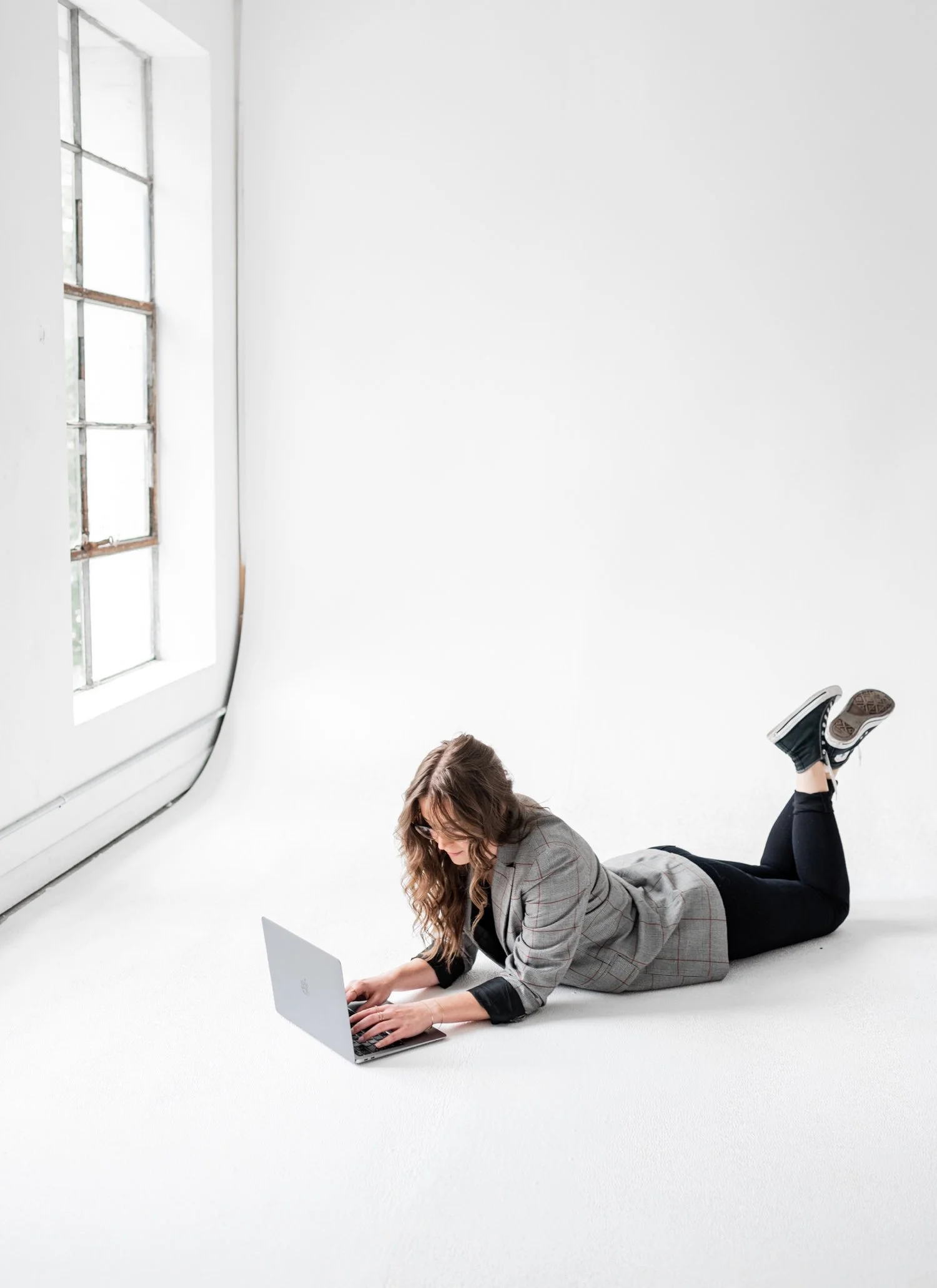 Woman lying on her stomach on the floor, working on a laptop in a bright room with white walls and large window.