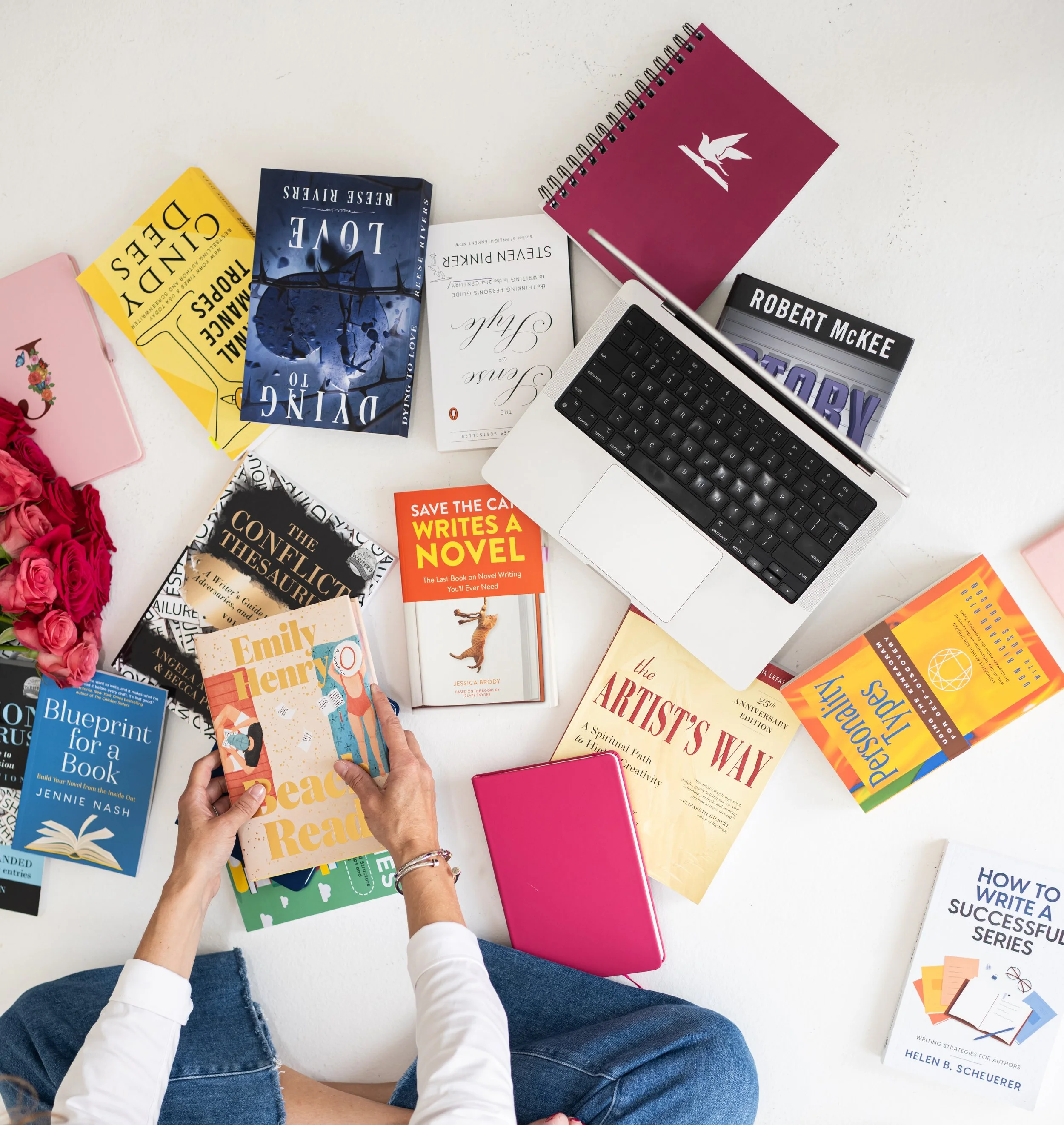 Person holding a book titled 'Beach Read' surrounded by various books, a laptop, notebooks, and roses on a white surface.