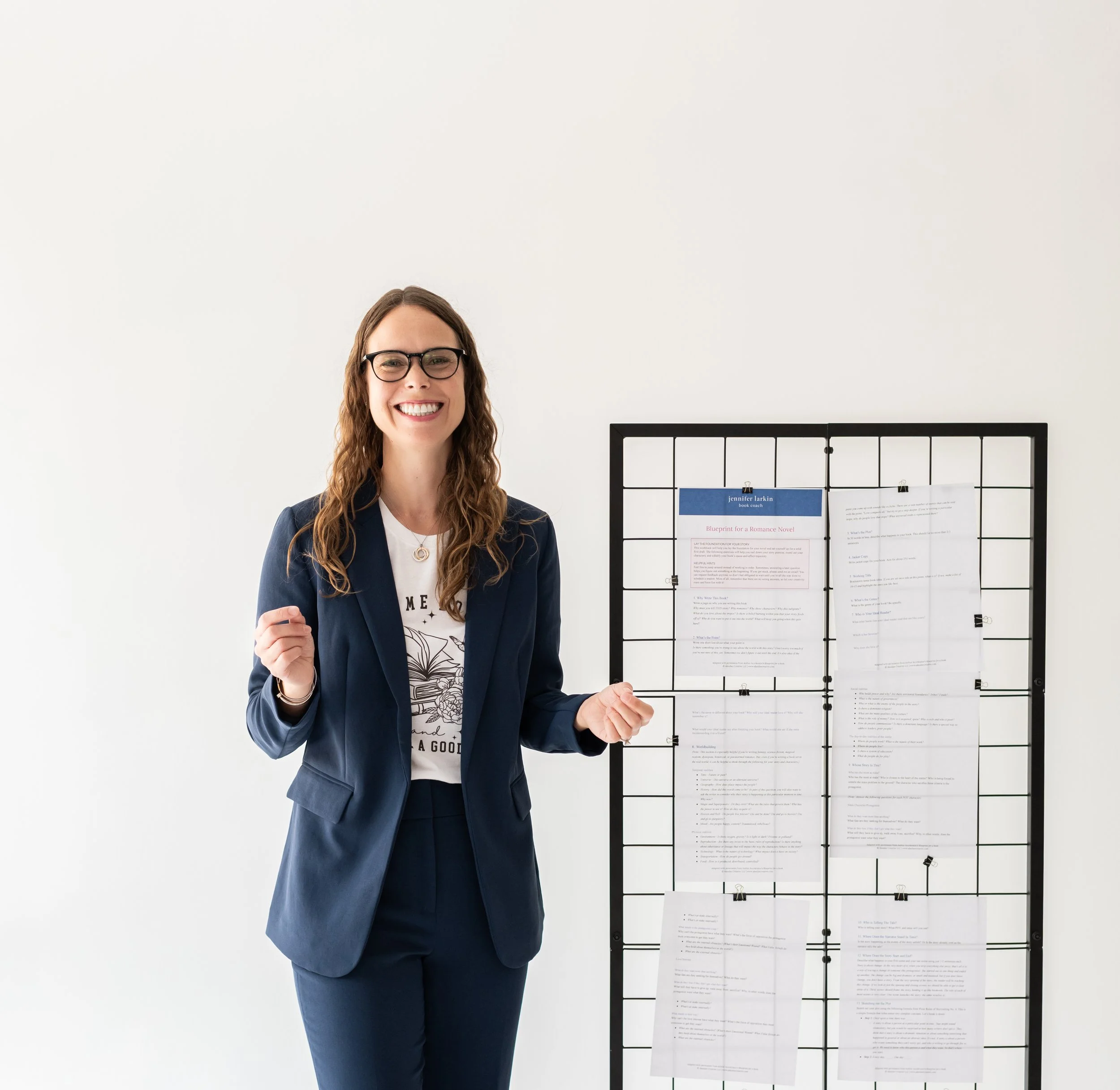 A woman in a navy blue suit standing next to a black wire grid with papers attached, smiling at the camera.