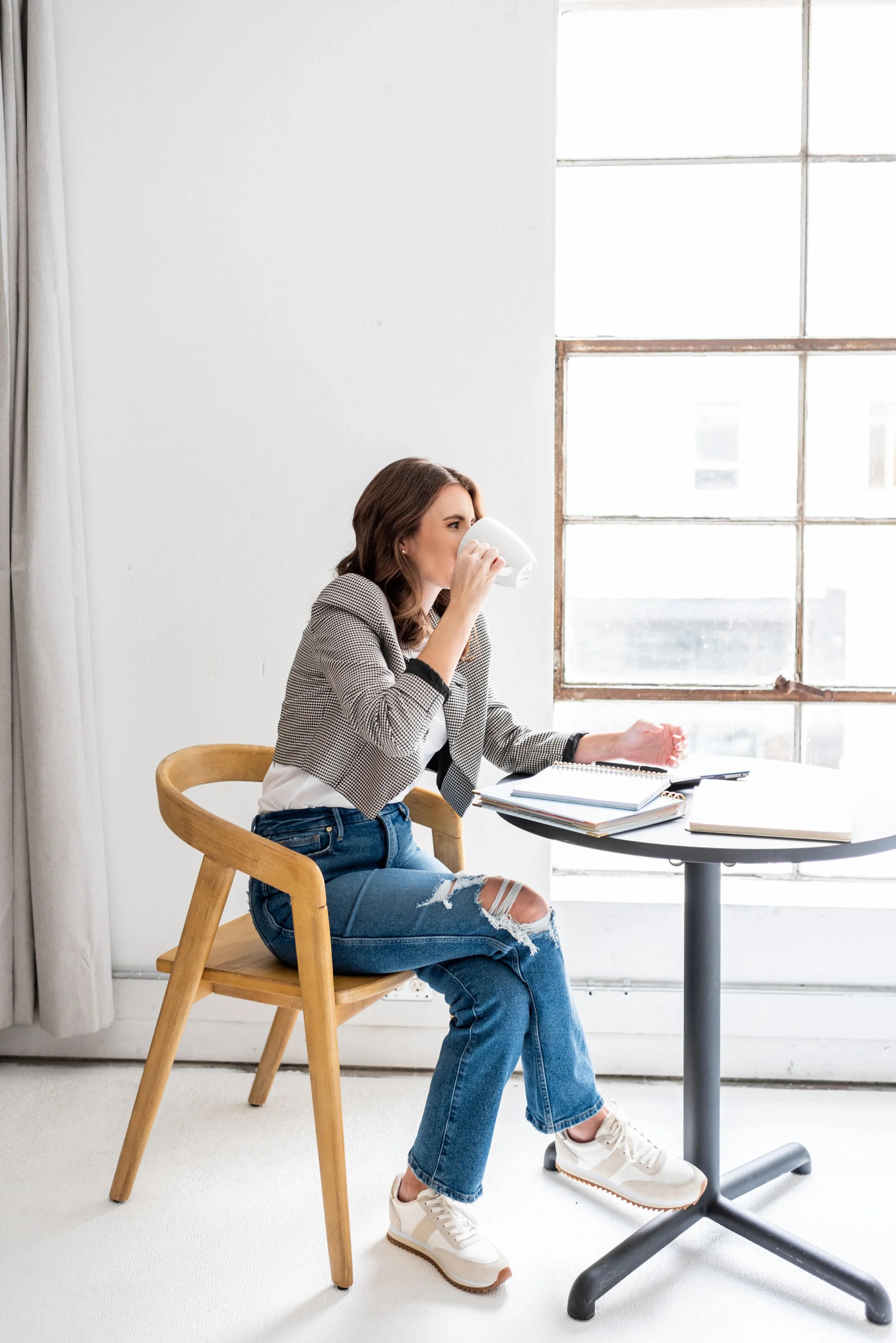 A woman sitting on a wooden chair, drinking coffee or tea from a white mug at a round black table with notebooks and a pen, next to a large window in a bright room.
