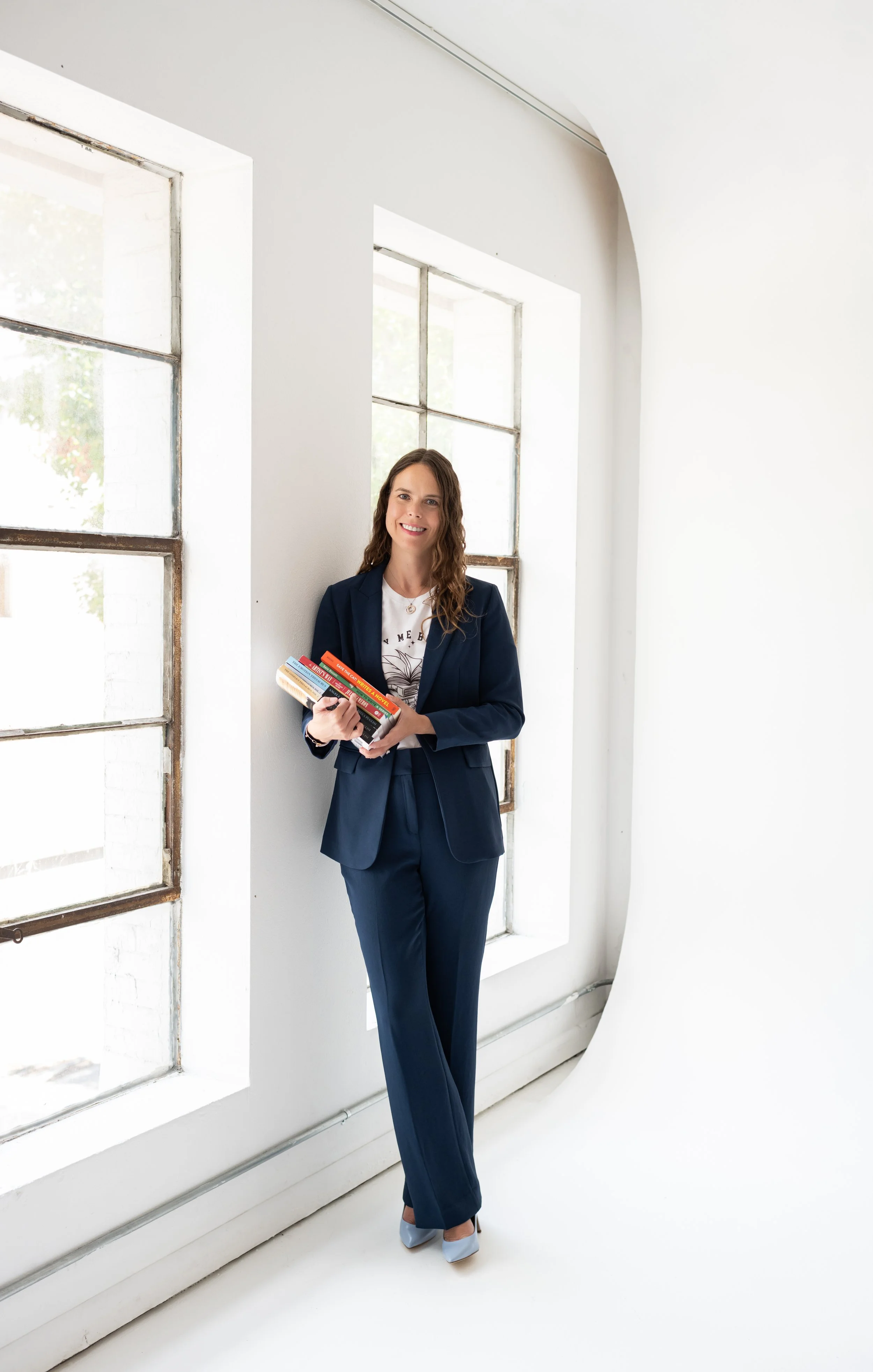 A woman in a navy blue suit holding a stack of books, standing in a bright room with large windows and white walls.