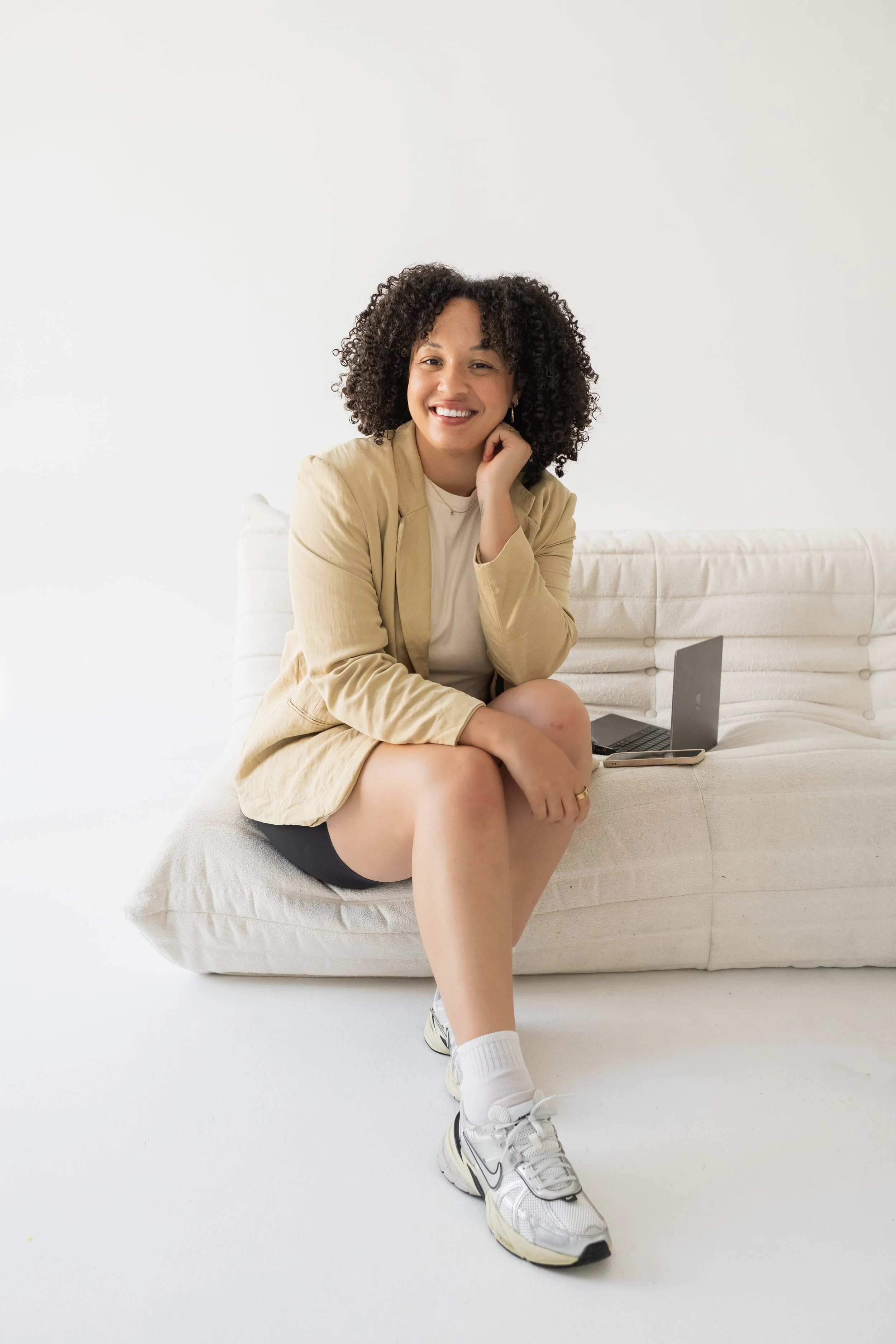 A woman with curly hair sitting on a white couch, smiling, with a laptop and smartphone beside her.