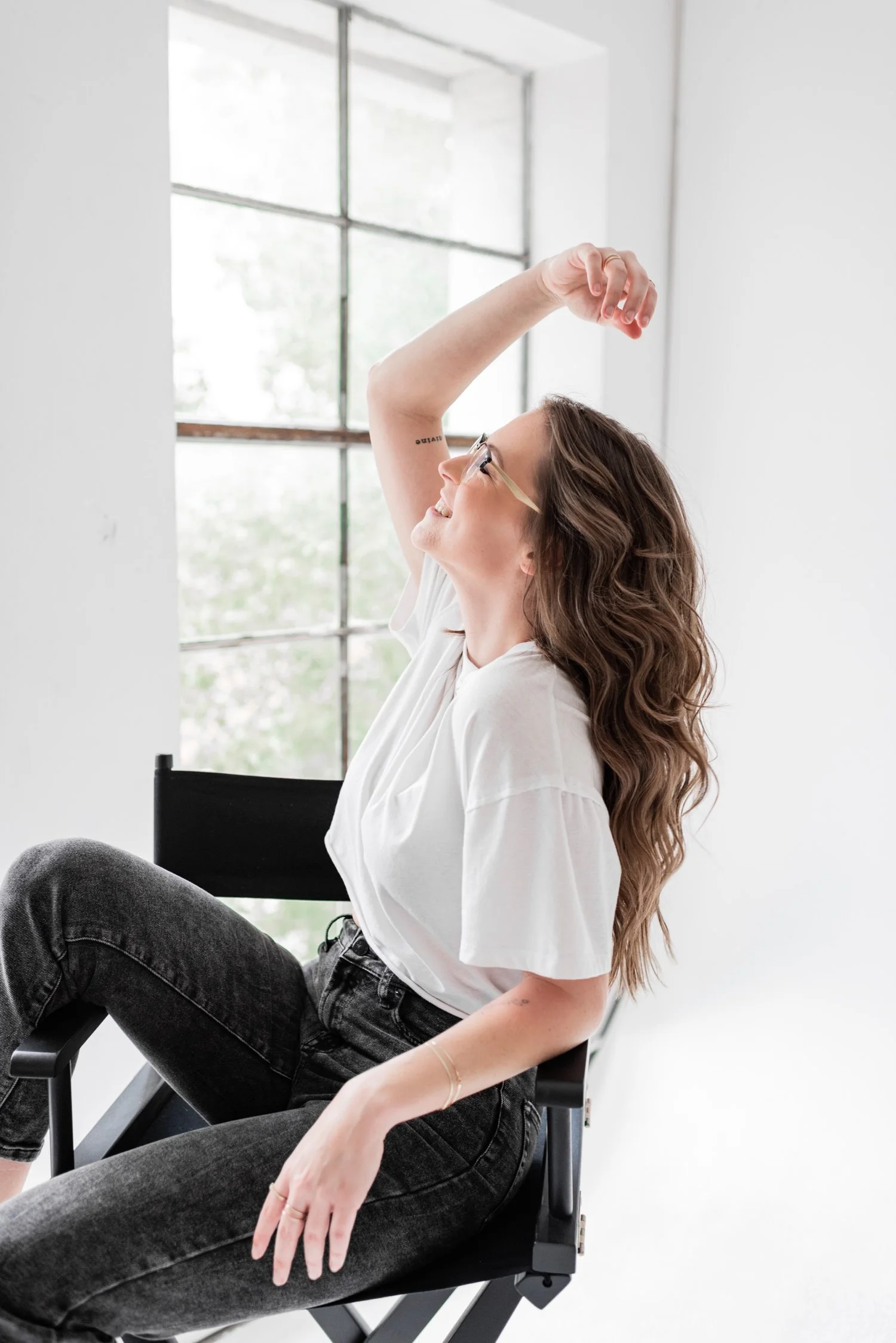 A woman sitting on a black chair near a large window, stretching her arm upward and looking up with a smile.