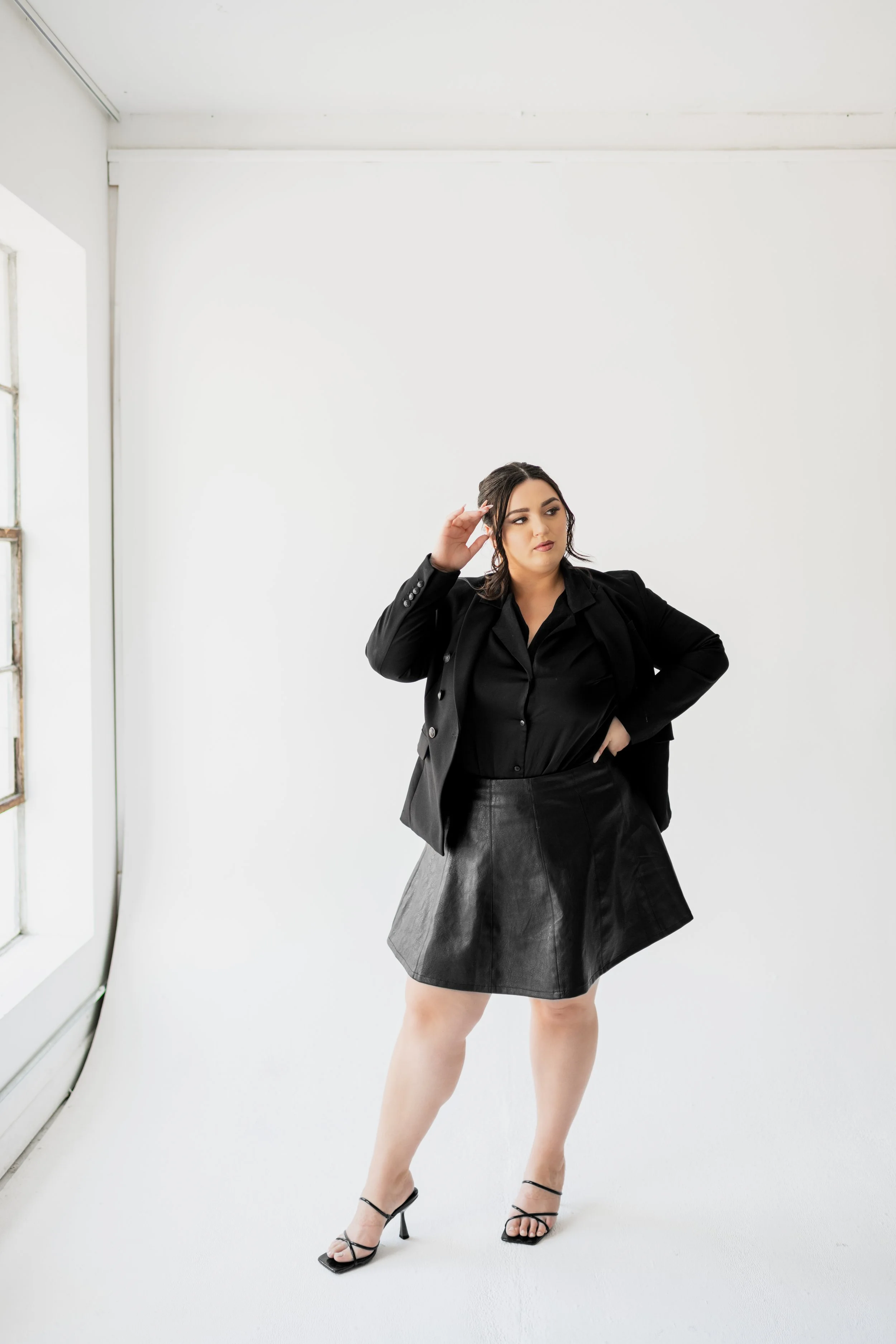 A woman with dark hair, wearing a black blazer, black blouse, leather skirt, and high-heeled sandals, standing in a bright white room near a window with her hand touching her head.