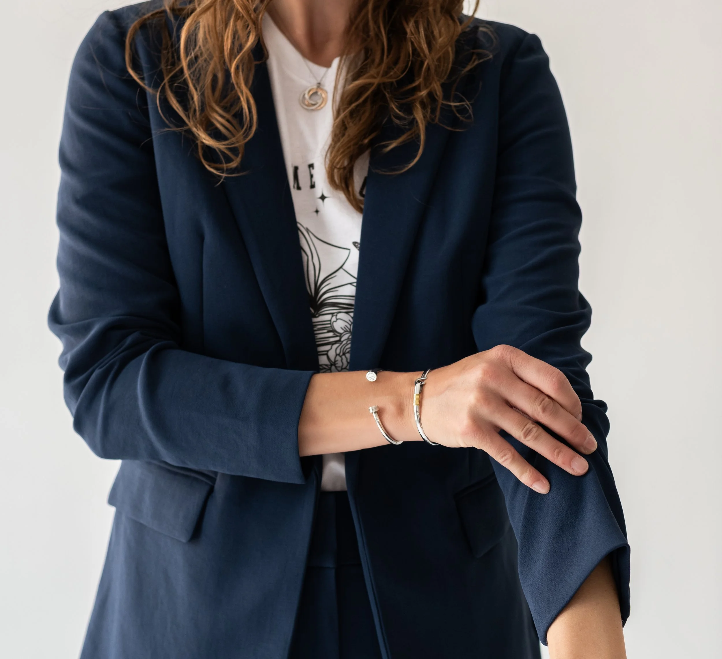 A woman with long wavy hair wearing a navy blazer over a graphic T-shirt and several bracelets, rolling up her blazer sleeve.