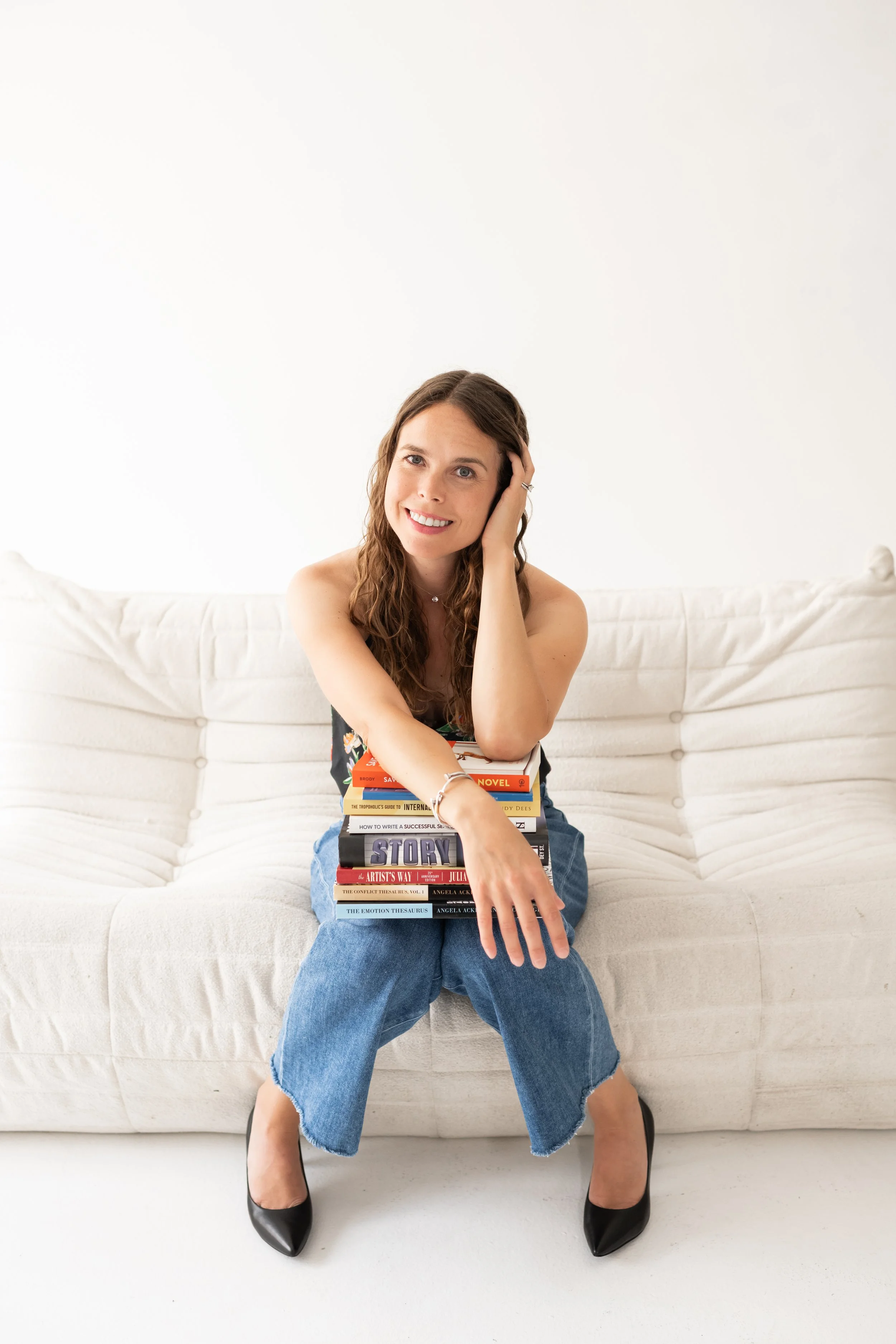 A woman sitting on a white sofa with a stack of books balanced on her lap, smiling and touching her head.