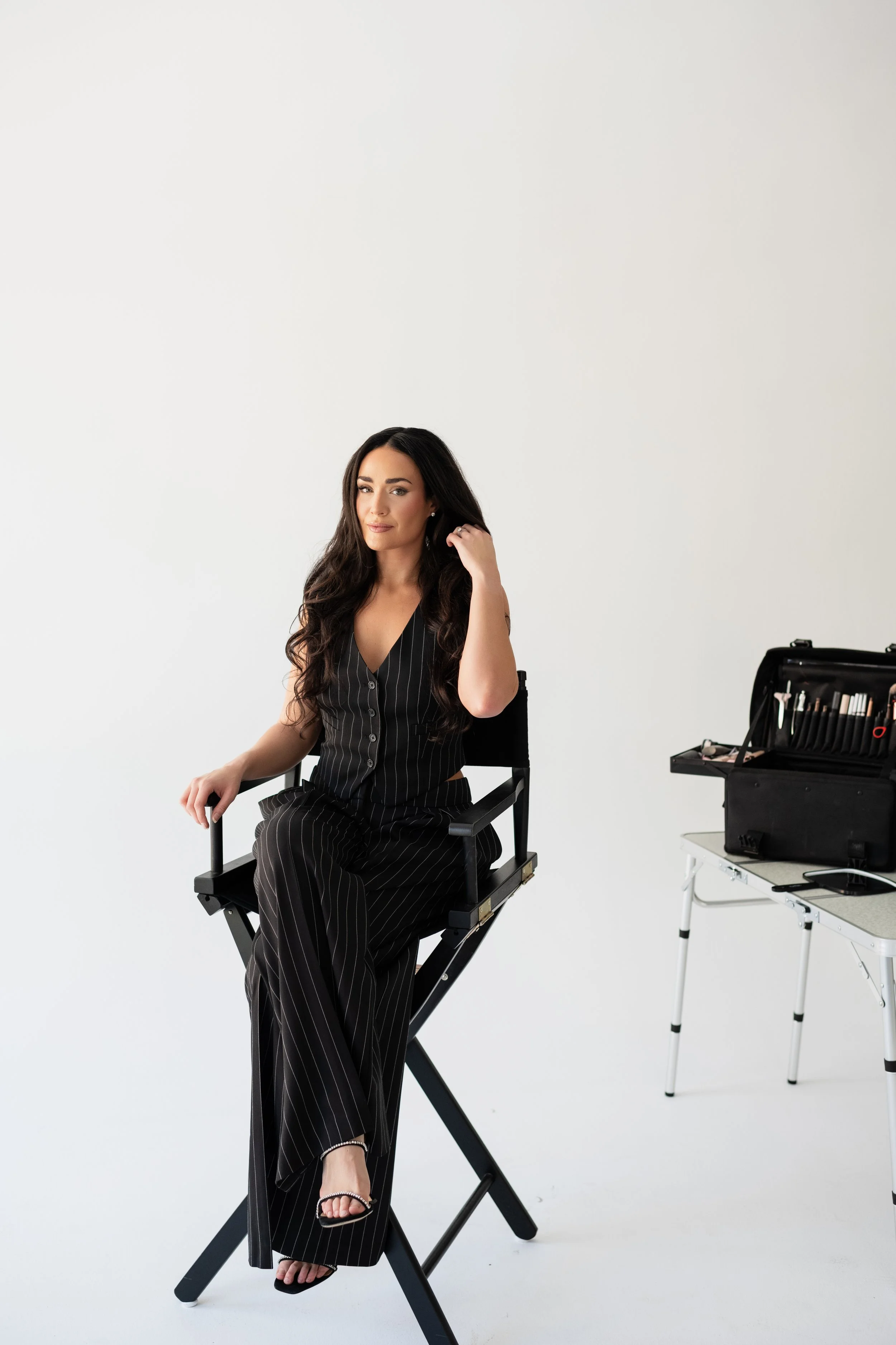 A woman with long dark hair, wearing a pinstripe suit, sitting on a director's chair in a photography studio, with makeup and beauty tools on a nearby table.