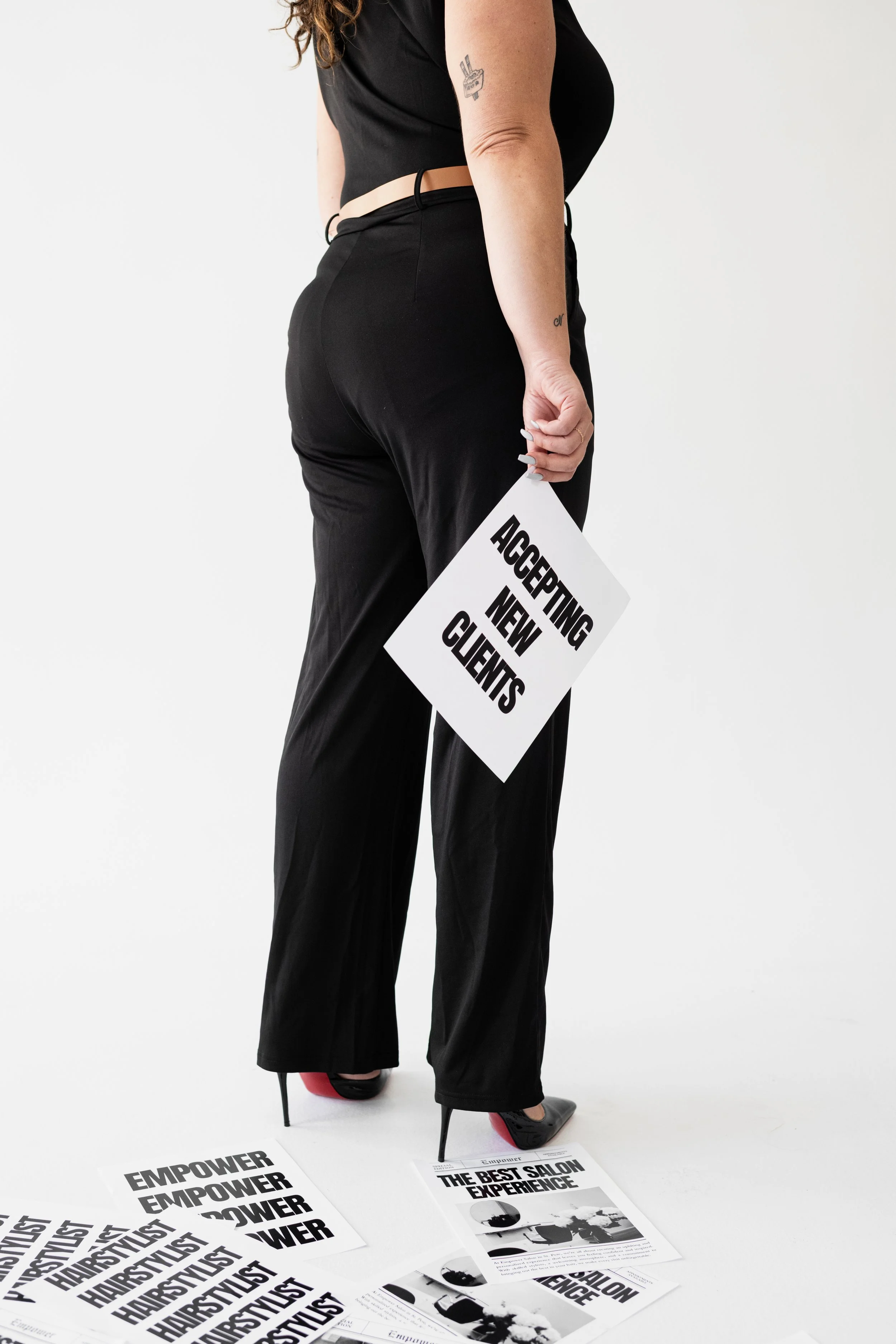 Woman standing with a sign that says "Accepting New Clients," surrounded by flyers on the floor, with a plain white background.