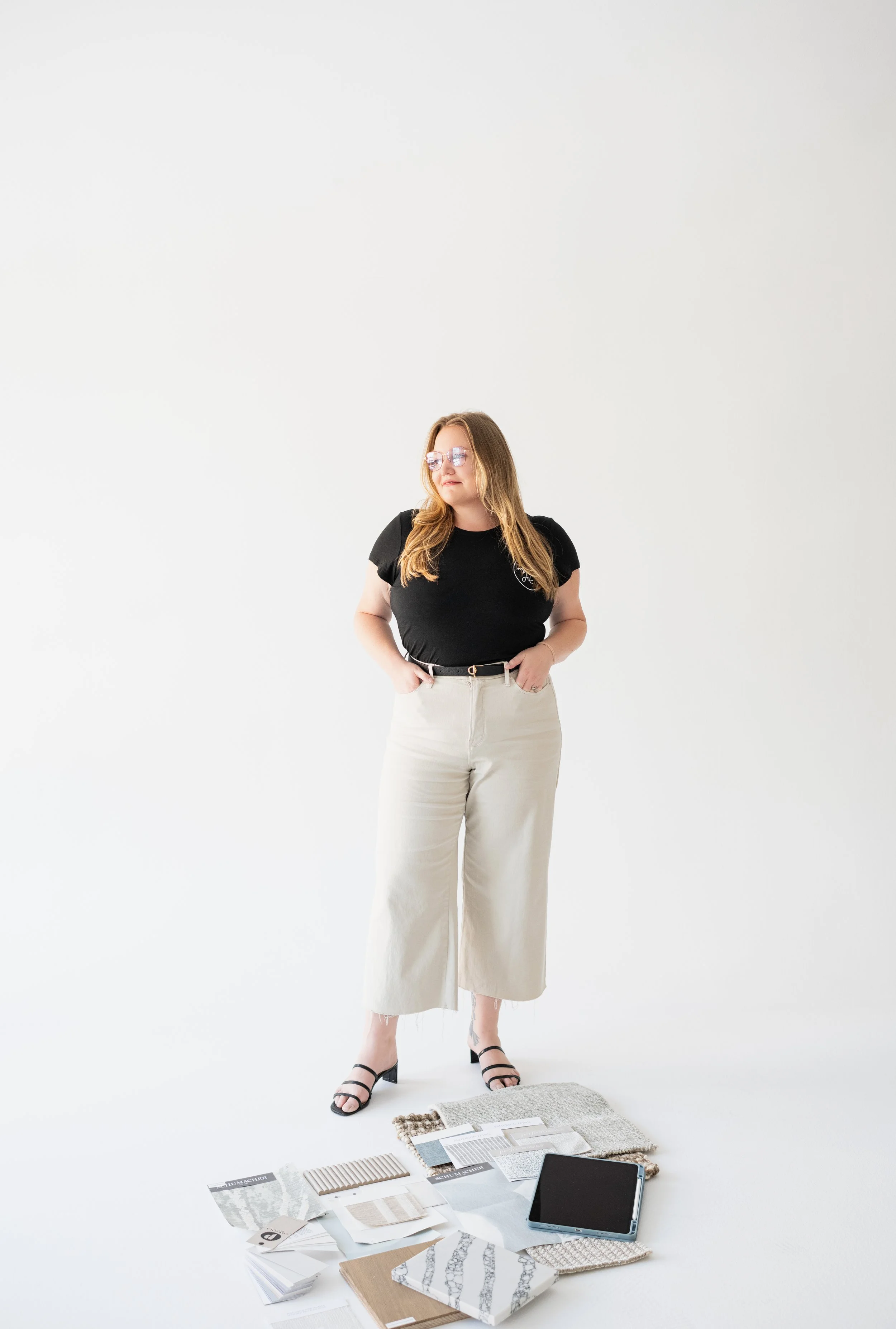 A woman wearing glasses, a black t-shirt, and beige wide-leg pants standing on a white background with various fabric samples, color swatches, and a tablet laid out in front of her.