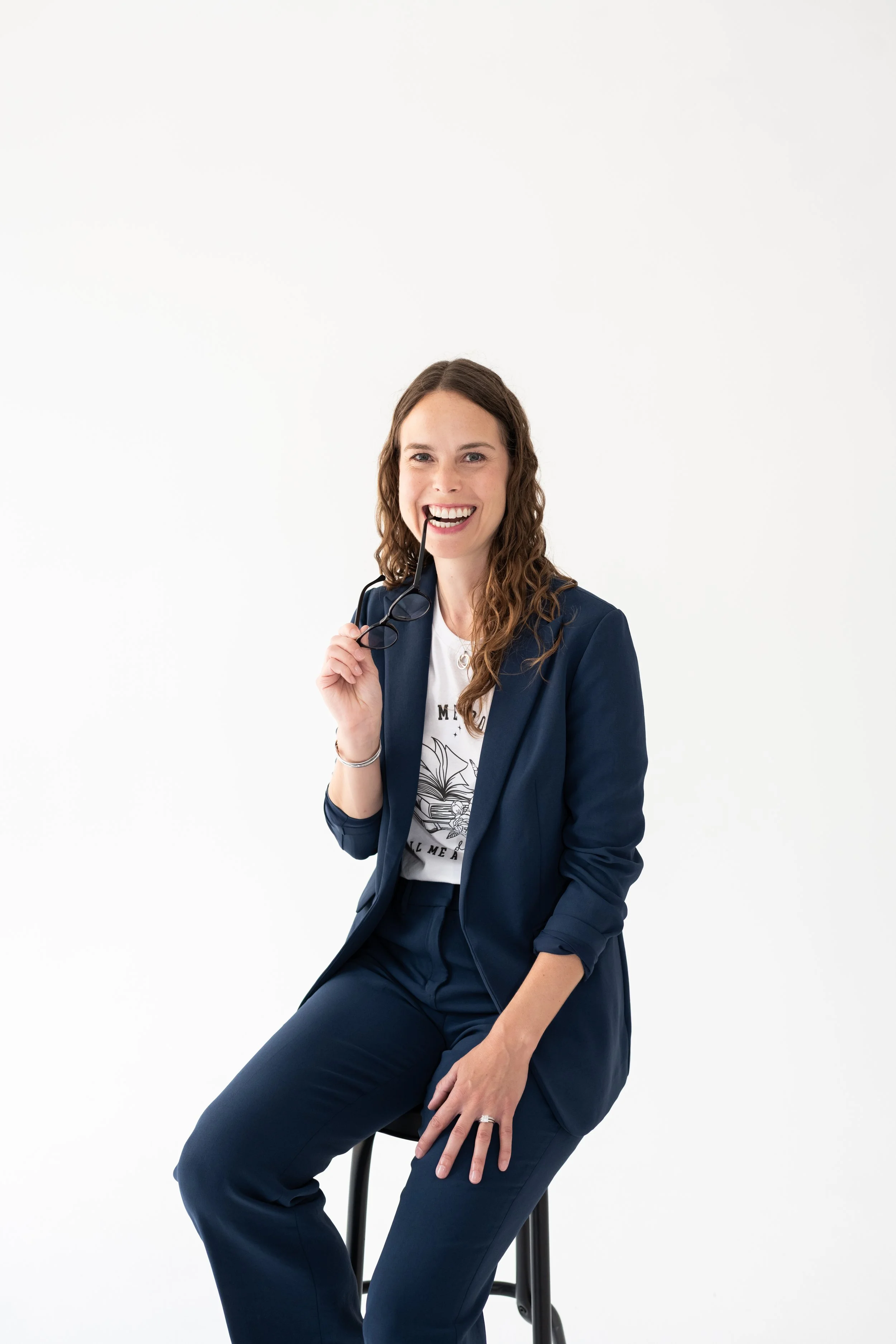 A woman in a navy blazer and pants, sitting on a stool, smiling, holding glasses with one hand, and biting the arm of her glasses, against a plain white background.