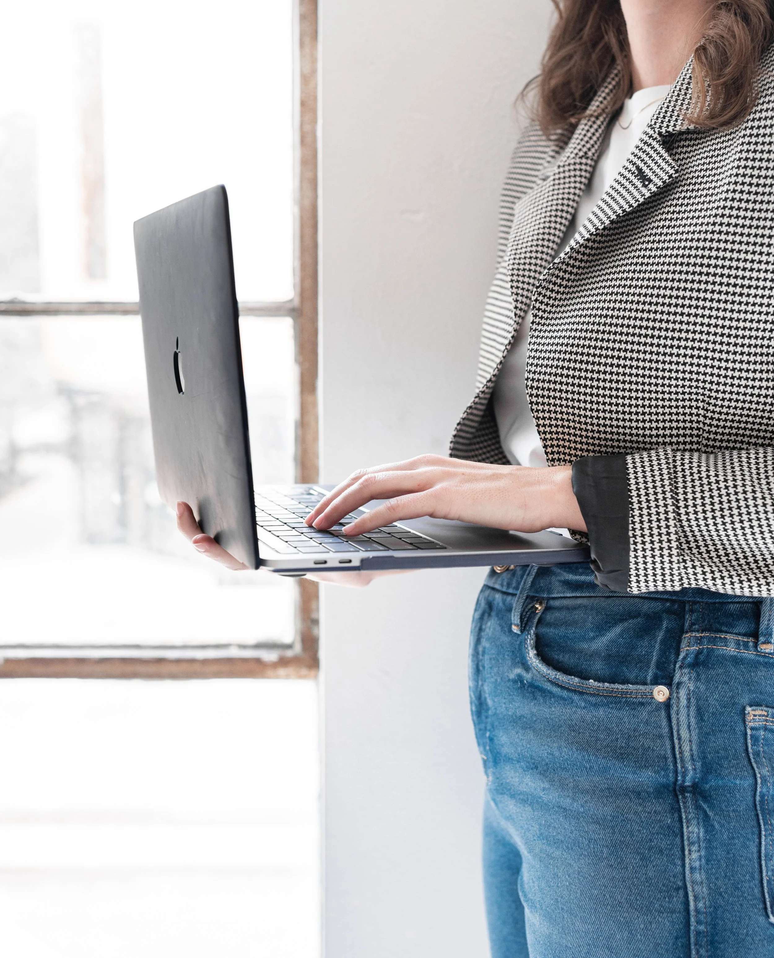 Person wearing jeans and a checkered blazer using a laptop near a window.