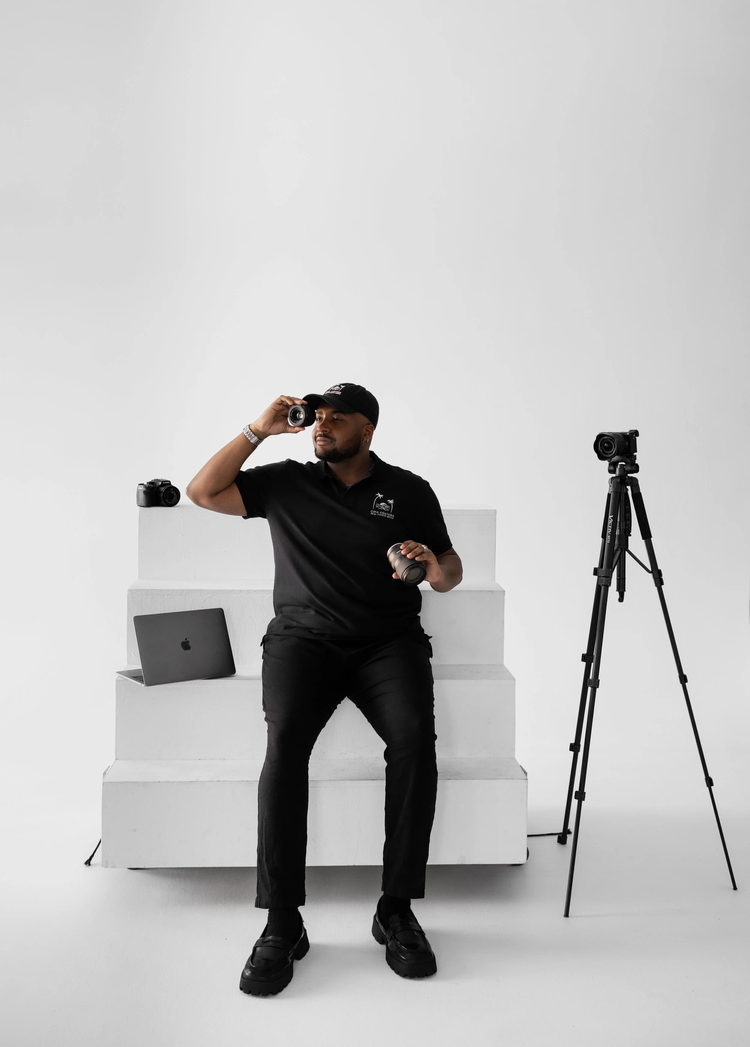 Photographer in black clothing and cap sitting on white steps, holding a camera to his eye, surrounded by photography equipment including a tripod, cameras, and a laptop, in a minimalist white studio.