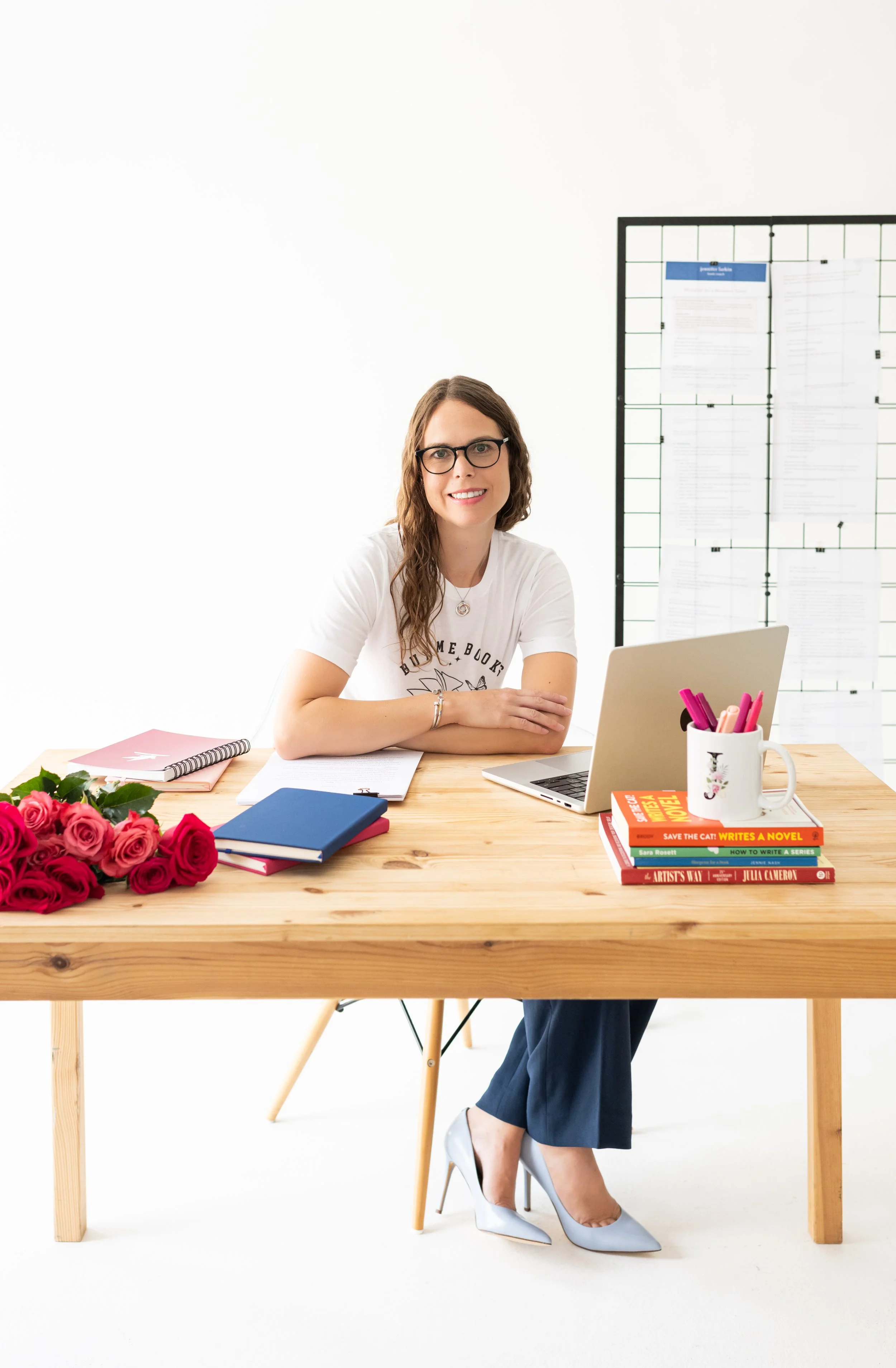 A woman with glasses and long brown hair sitting at a wooden desk with a laptop, pink roses, notebooks, and books, in a bright room with a white background and a black wire grid in the background.