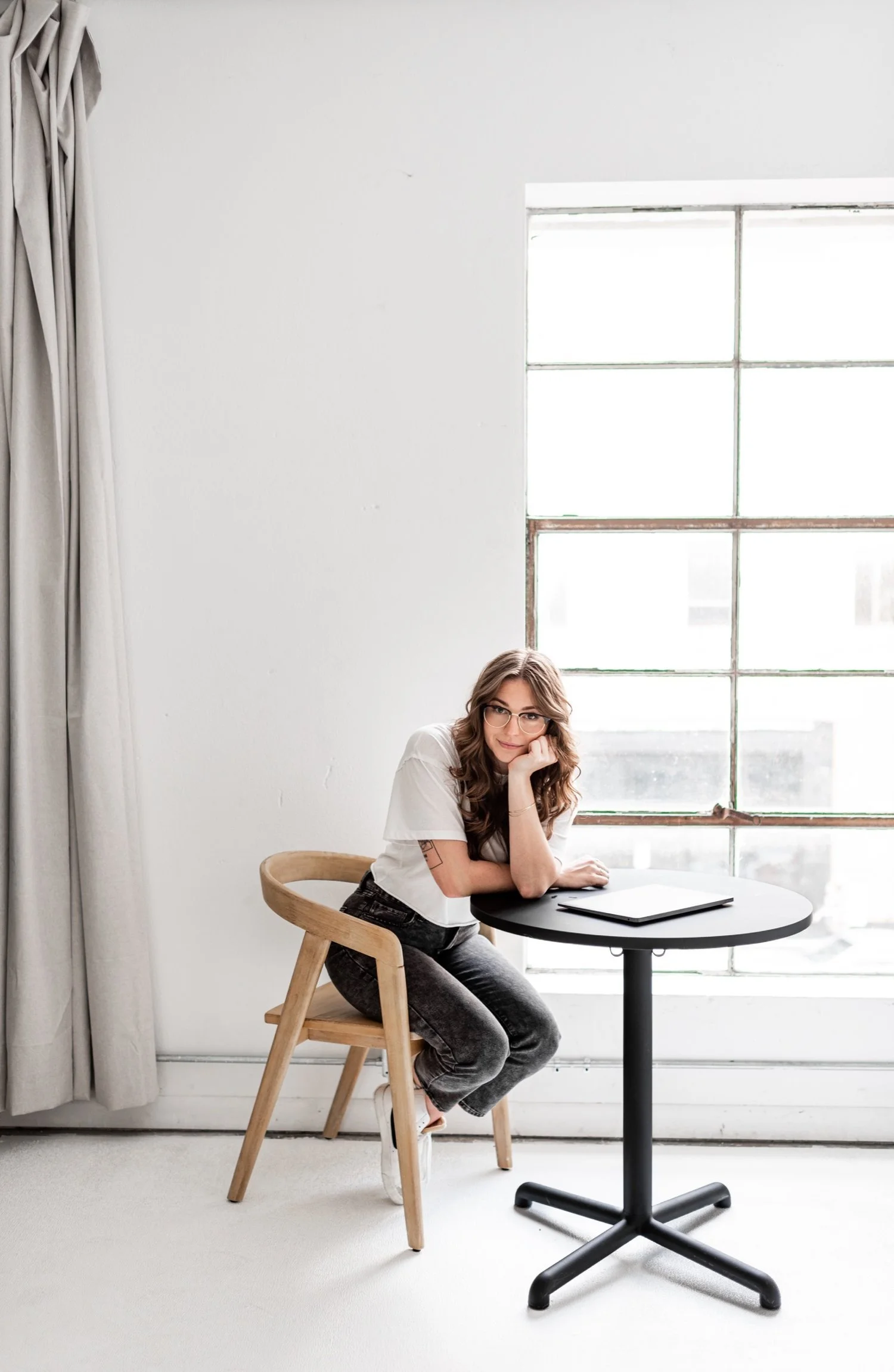 A young woman with wavy brown hair and glasses sitting at a small black round table with a closed laptop on it, resting her chin on her hand and looking at the camera, in a bright room with a large window, white walls, beige curtains, and a light uph