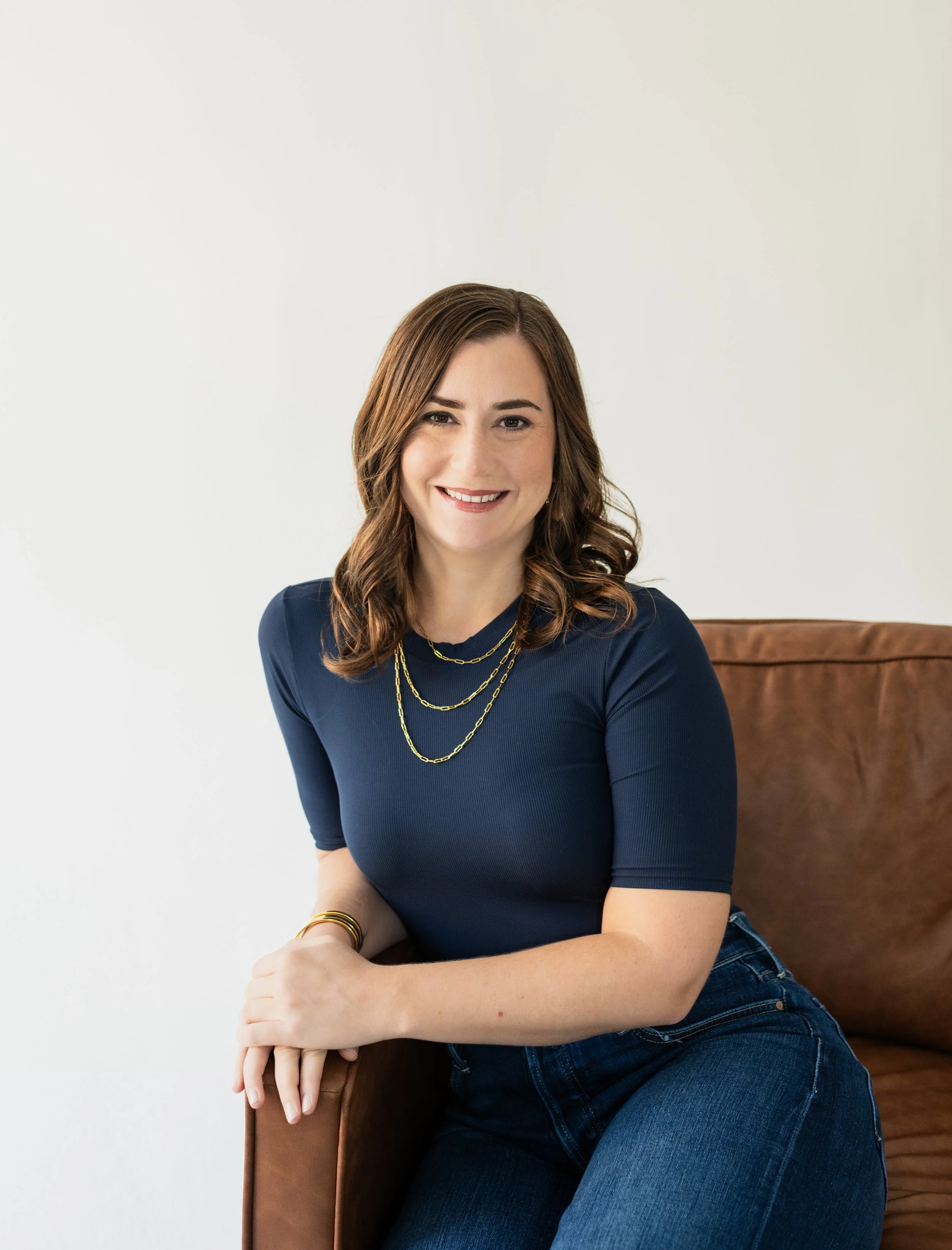 A woman with shoulder-length brown hair, smiling, wearing a navy blue top and layered gold necklaces, sitting casually on a brown leather chair against a plain white background.