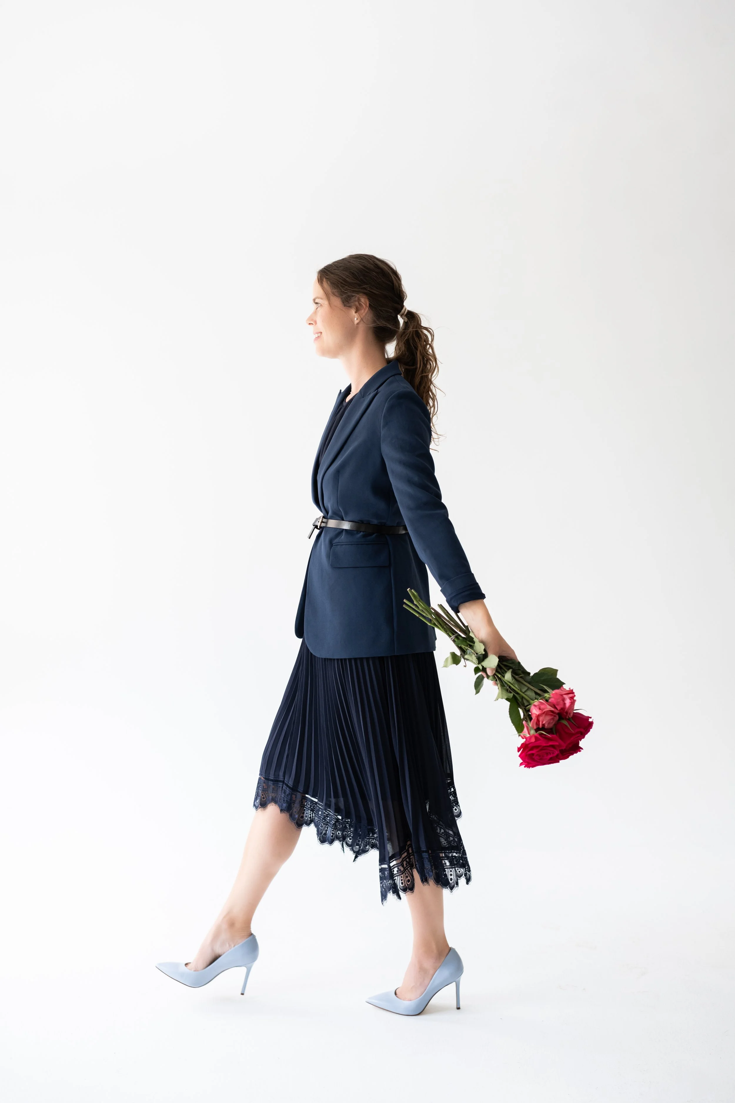 Woman in navy blazer, pleated lace skirt, and high heels walking with pink roses behind her back against a white background.