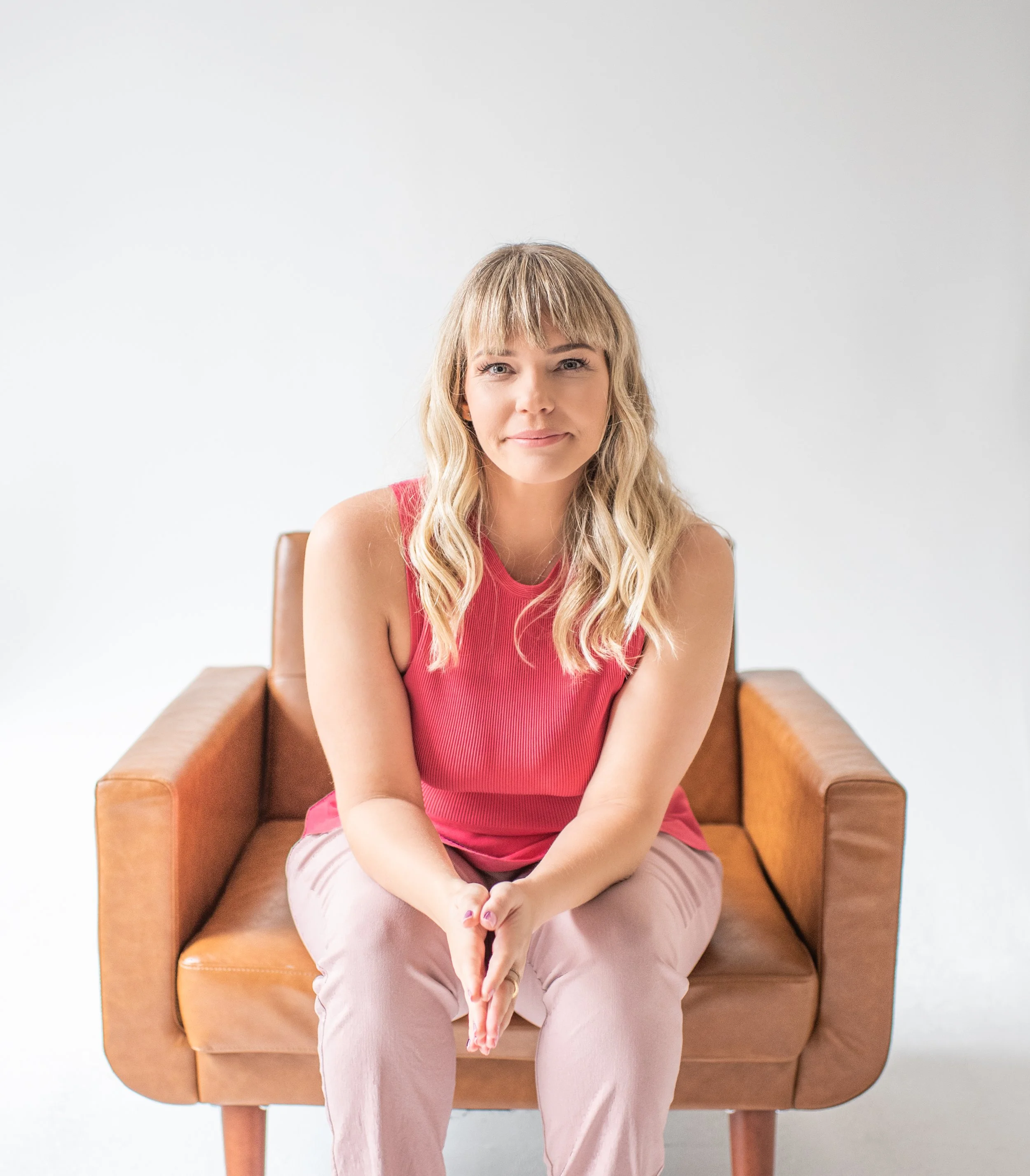 A woman with wavy blonde hair sitting on a tan armchair, wearing a sleeveless pink top and light pink pants, looking at the camera with a slight smile against a plain white background.