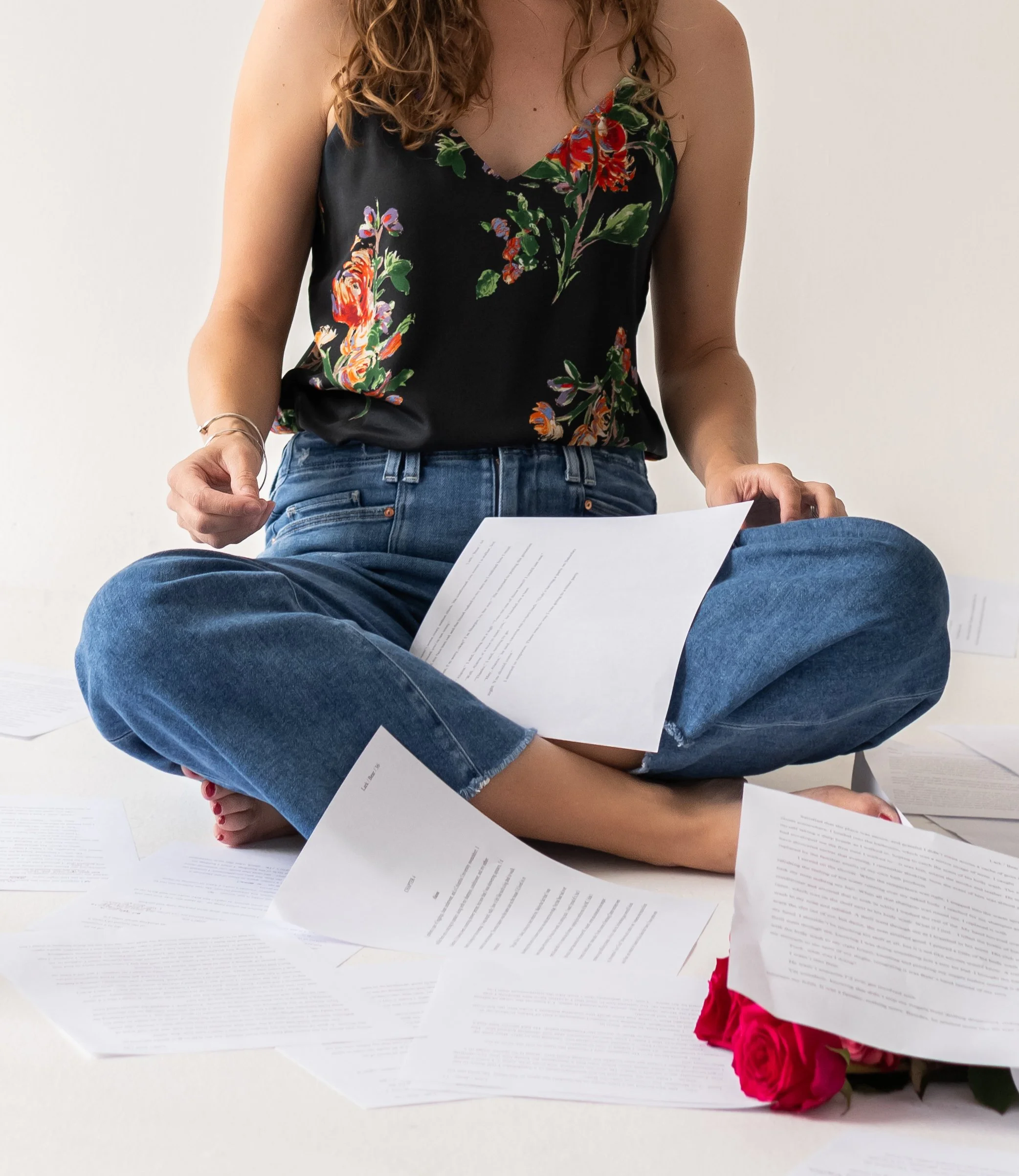 A woman sitting cross-legged on the floor with scattered papers and a bouquet of pink roses nearby, wearing a floral sleeveless top and blue jeans.