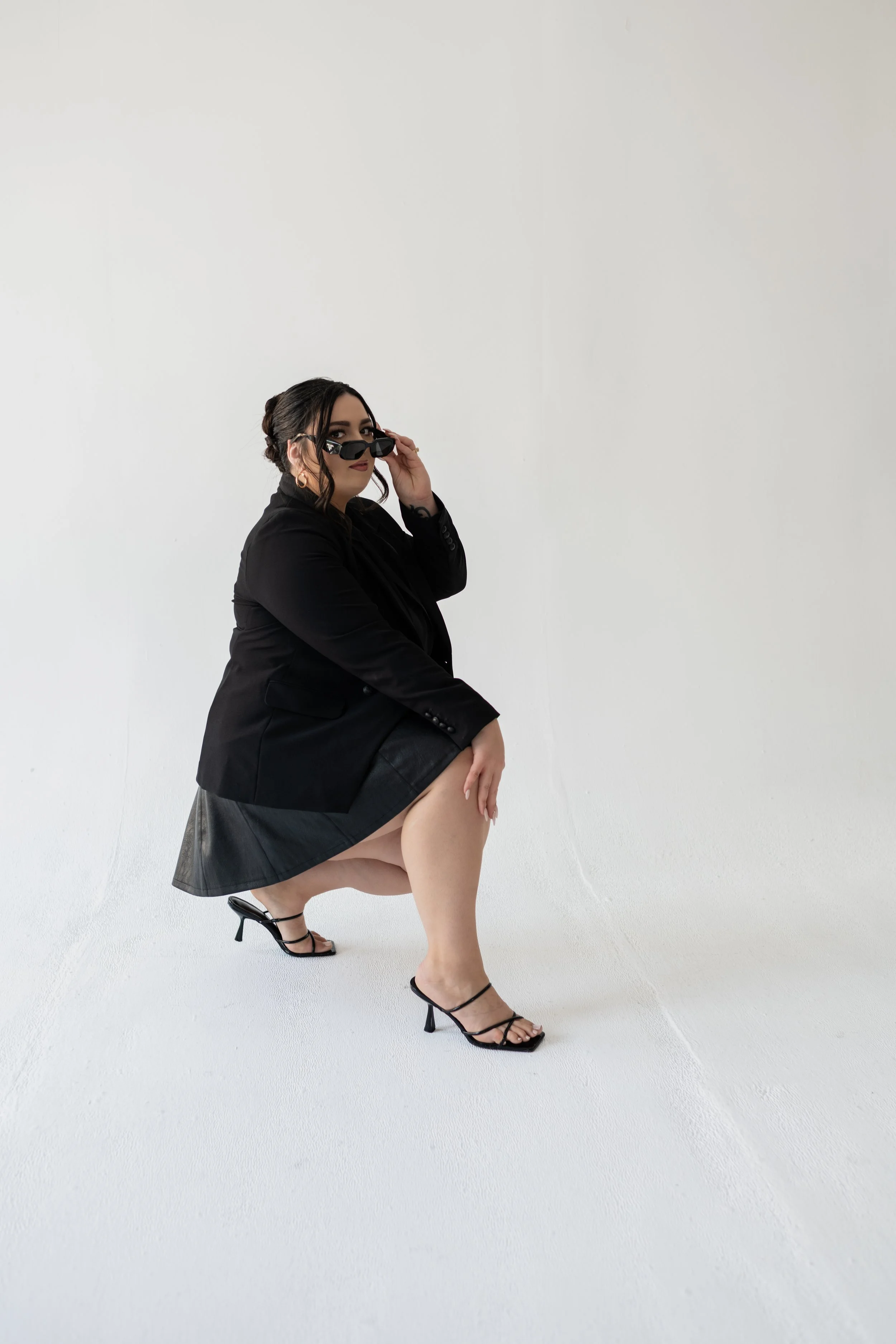 Woman in black blazer, black heels, and sunglasses squatting on a white floor and wall, adjusting her sunglasses.