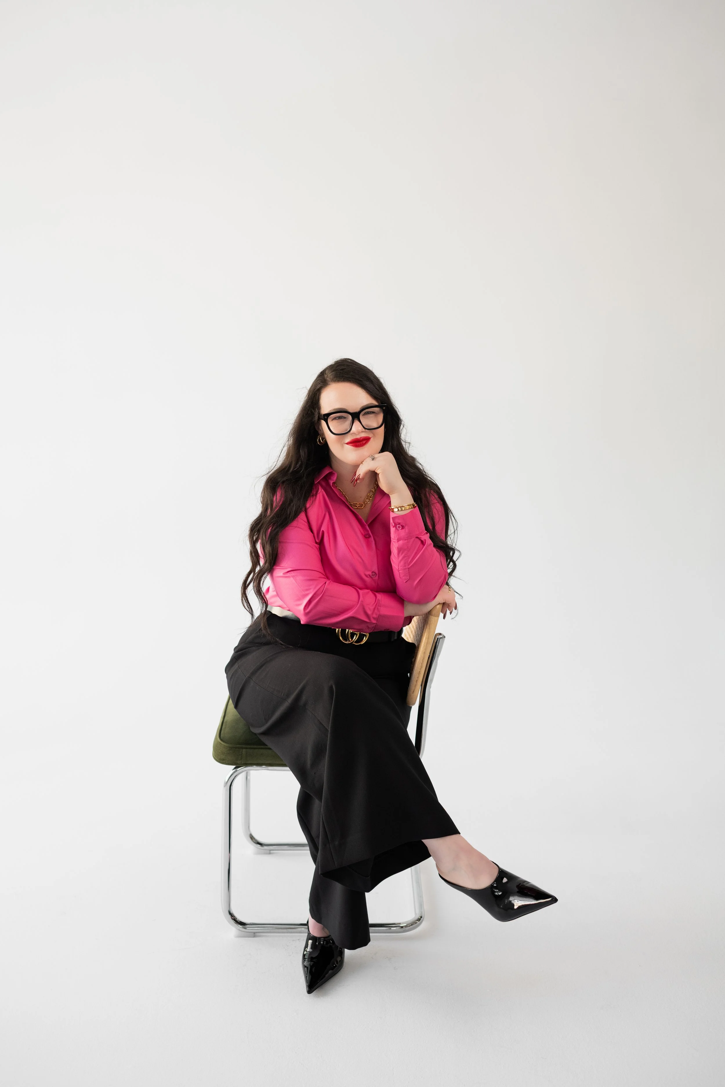 A woman with long dark hair, black glasses, red lipstick, wearing a bright pink blouse, black pants, and black shiny heels, sitting on a chair with a wooden backrest, against a plain white background.