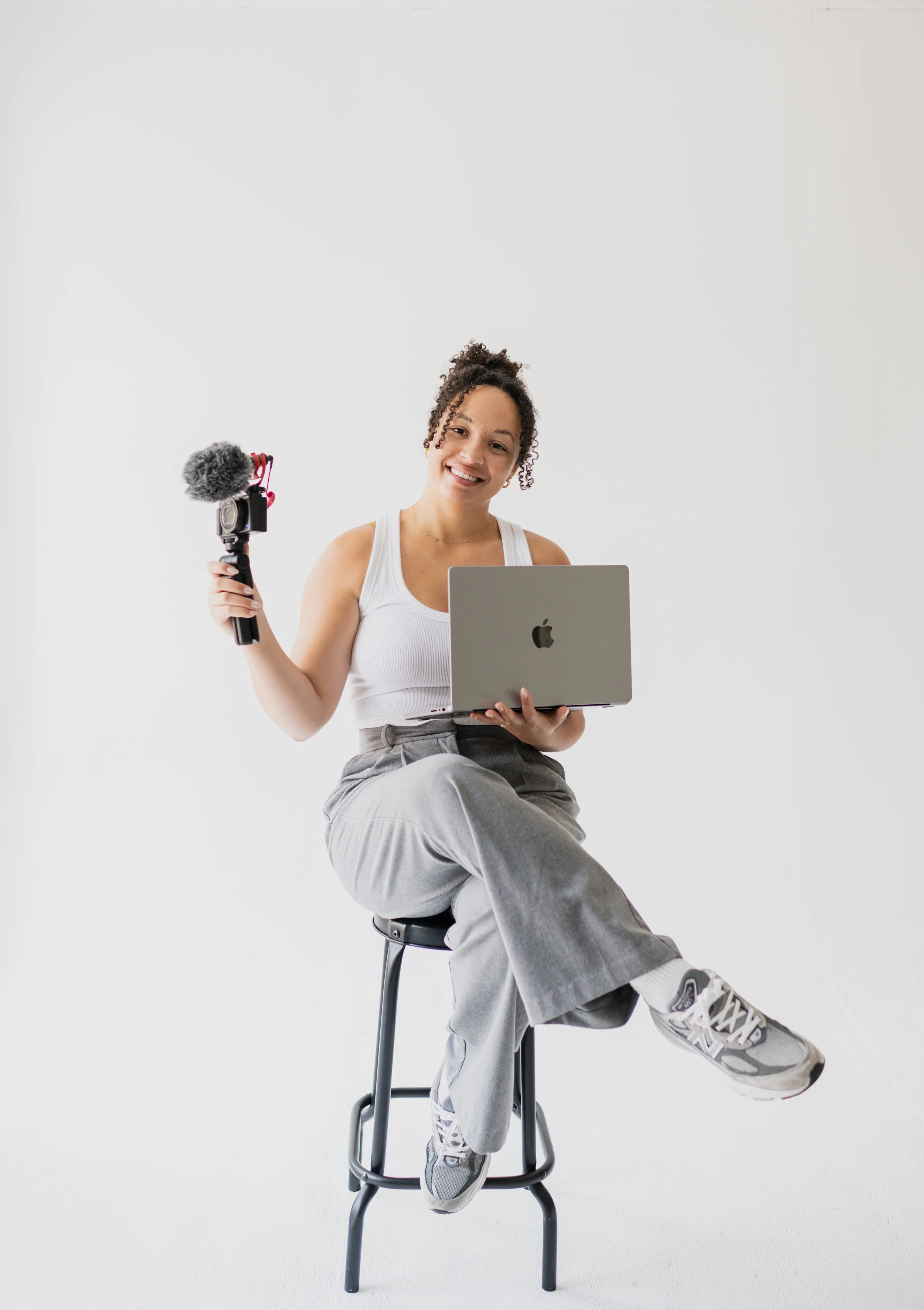 Young woman sitting on a stool with crossed legs, holding a laptop in one hand and a camera with a microphone in the other, smiling against a white background.