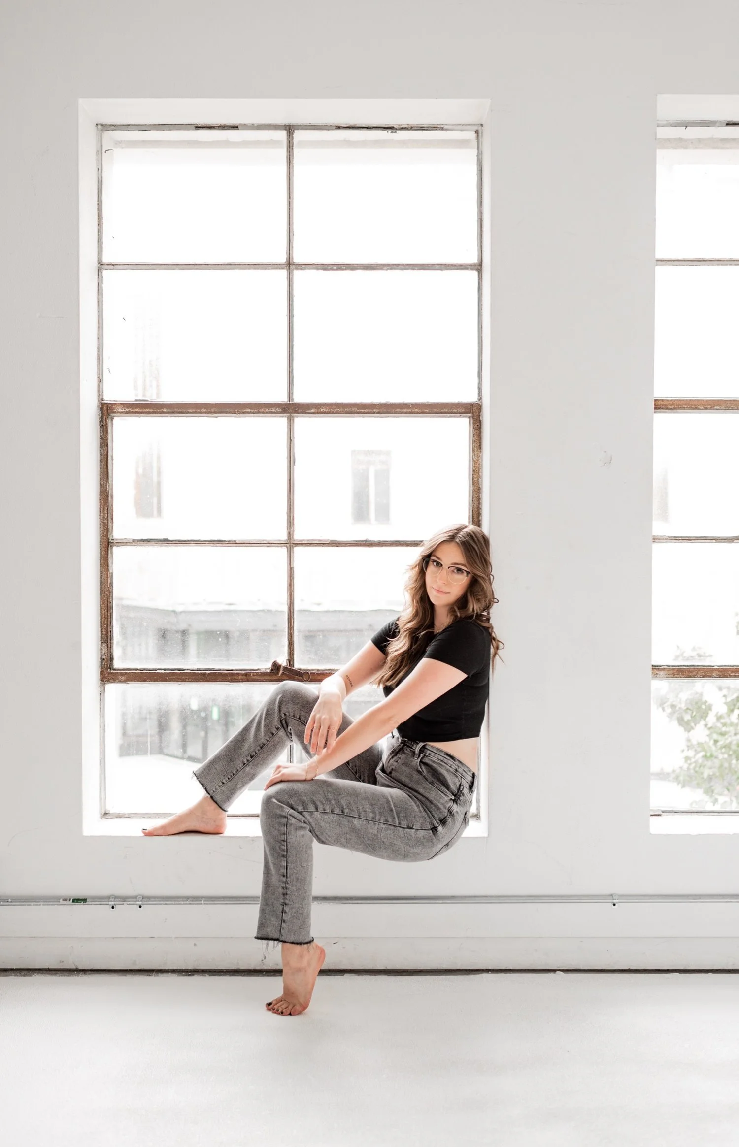 A woman sitting on a wide windowsill in a bright room with large grid windows. She is wearing glasses, a black crop top, and gray jeans, with one foot barefoot on the white floor.