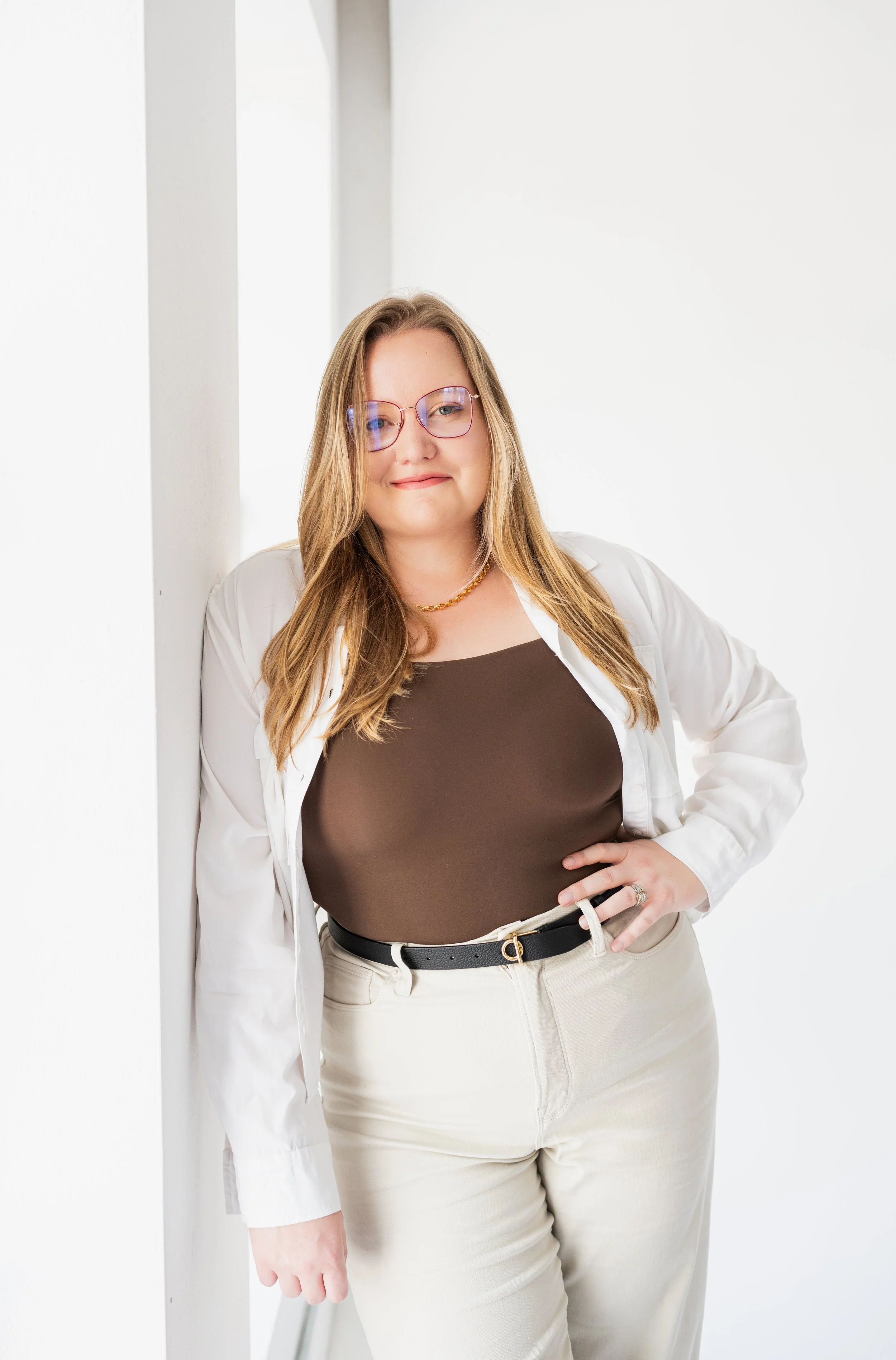 A woman with long blonde hair wearing glasses, a white open shirt, brown top, beige pants, and a black belt, standing against a white wall.