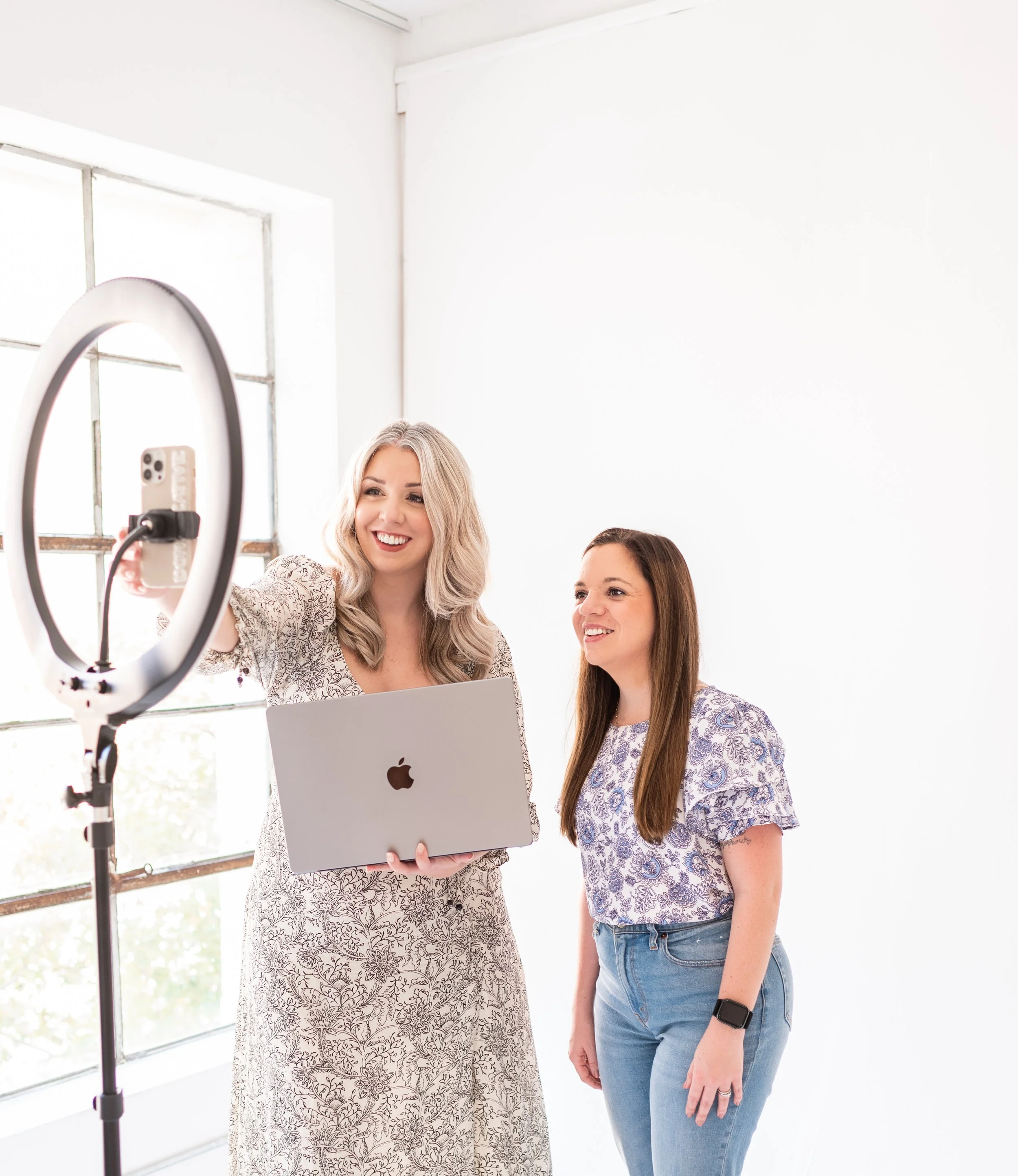 Two women taking a selfie using a ring light and a smartphone, one holding a laptop, both smiling in a bright room with white walls and a large window.